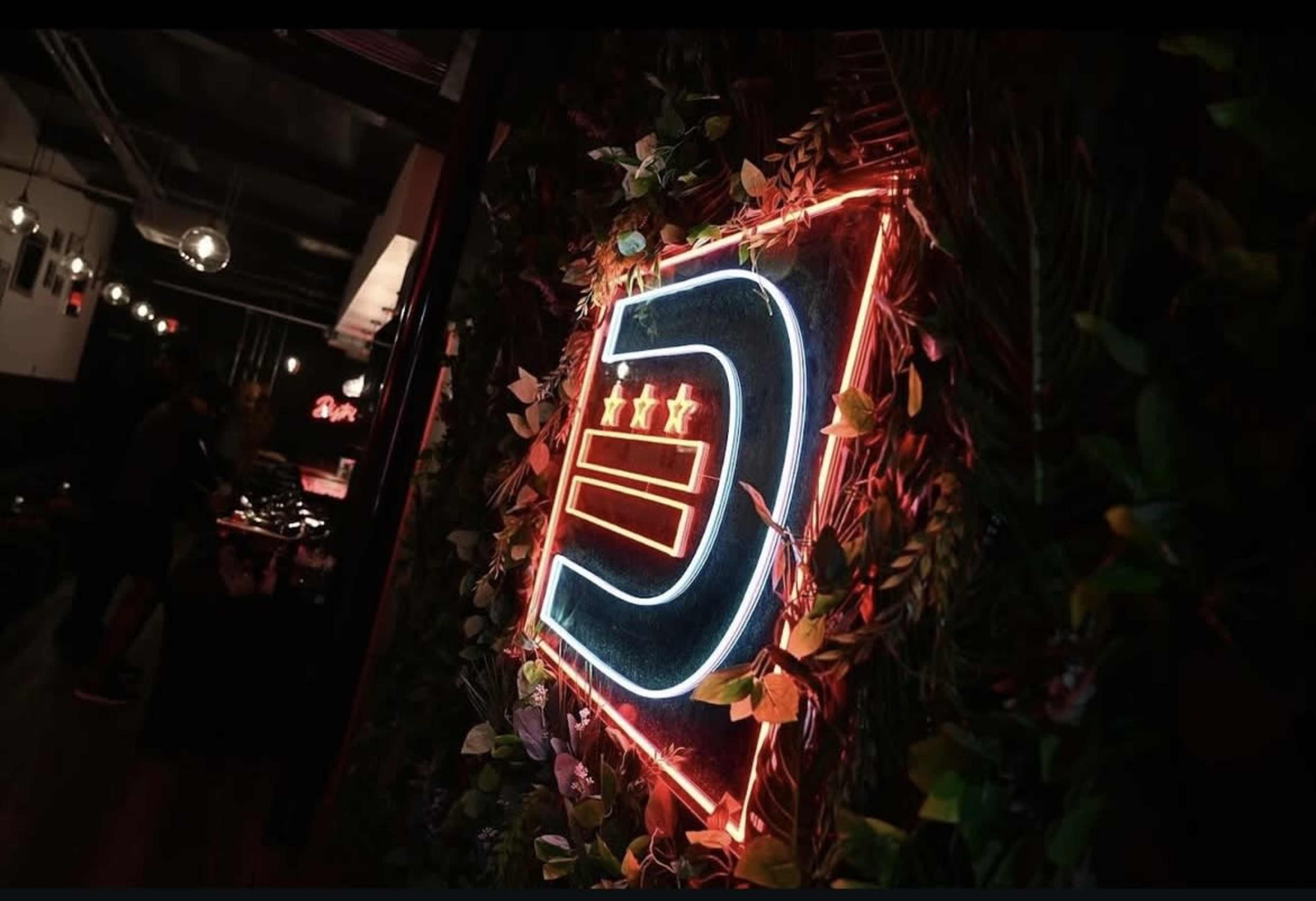 The image features a brightly lit neon sign with a stylized letter "D" surrounded by greenery in an indoor setting.
