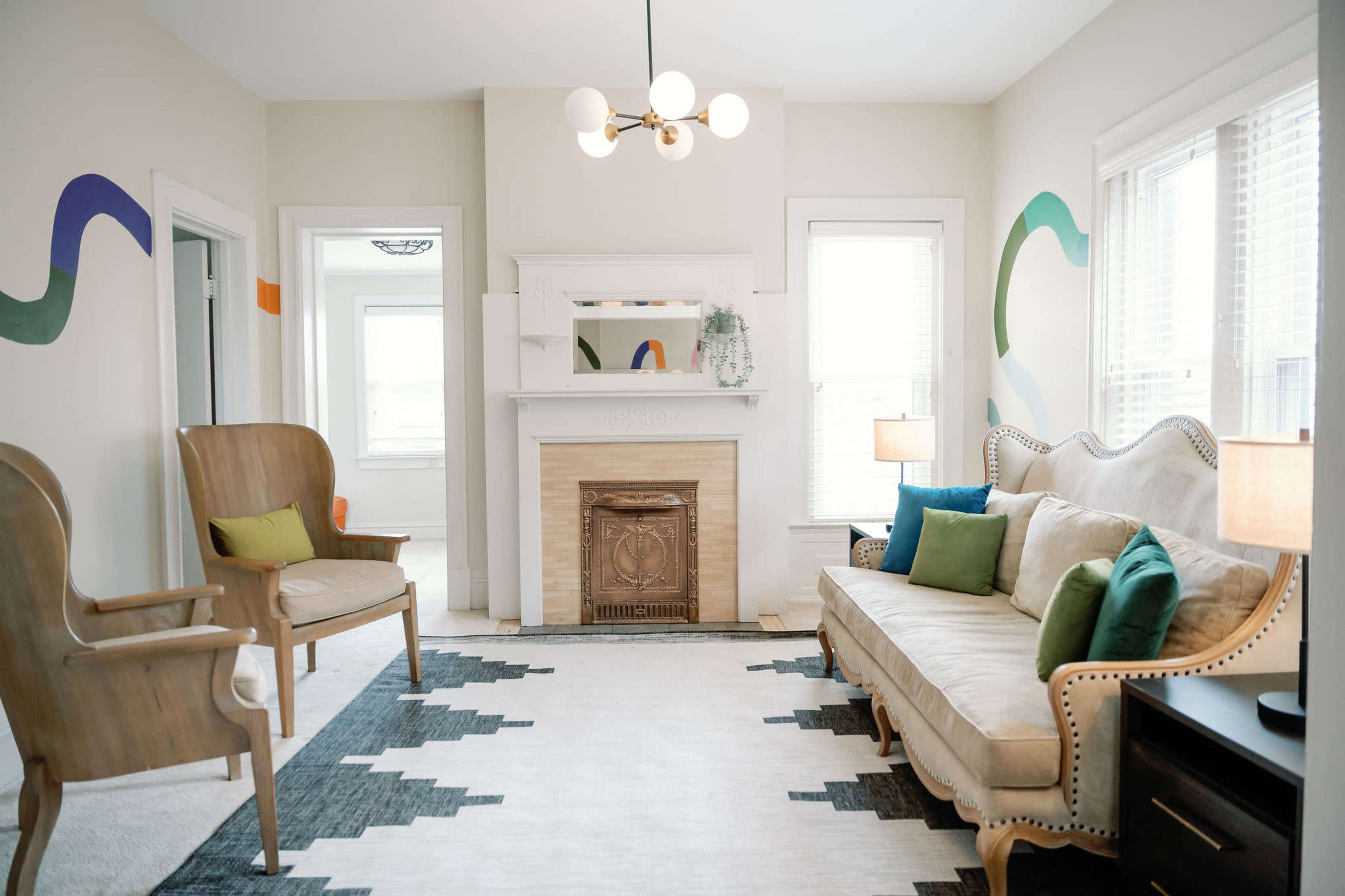 A living room with a beige sofa, two wooden armchairs, a fireplace with a mantle, and a patterned rug.