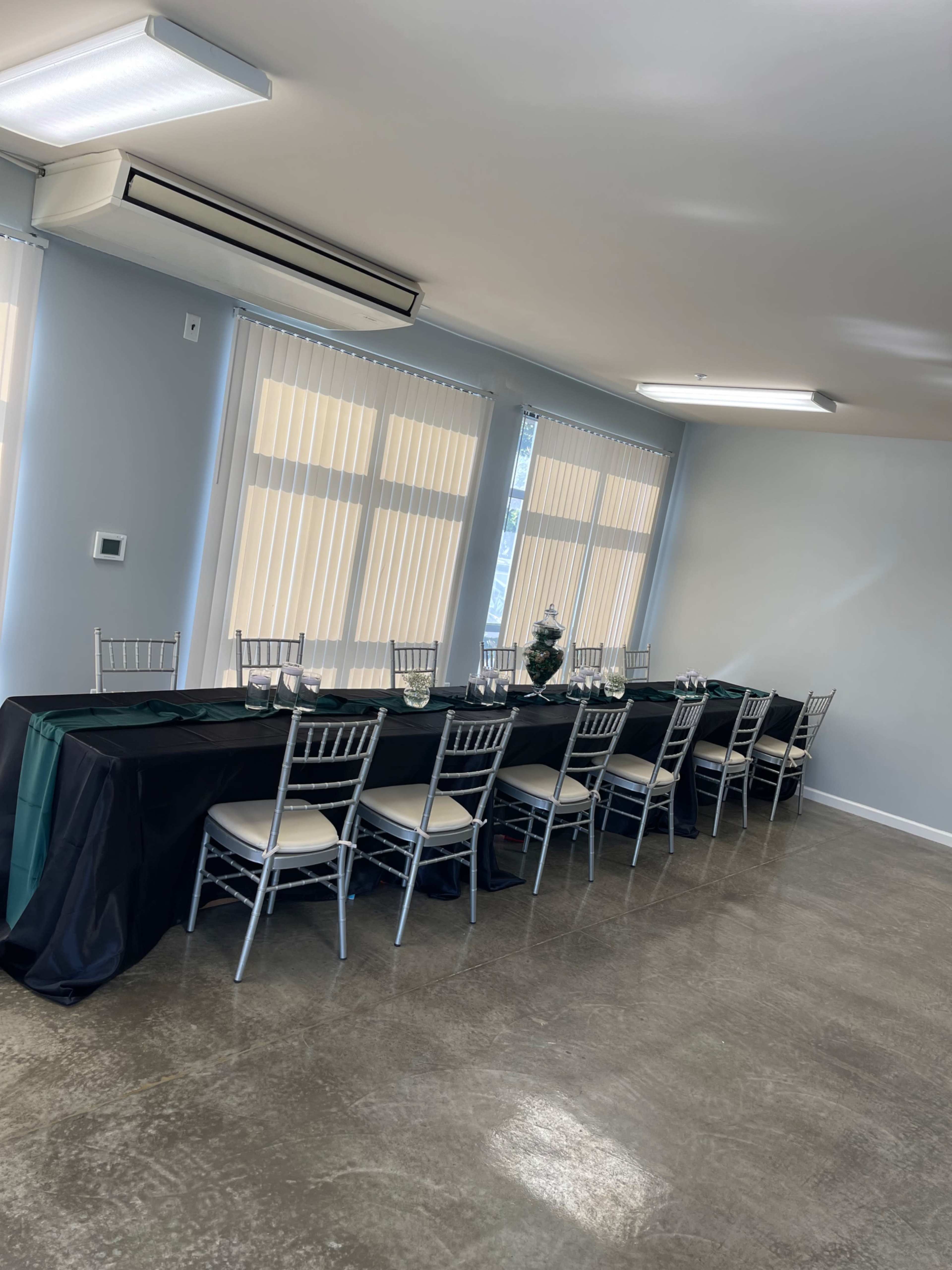 A long dining table with silver chairs is set against a wall of large windows in a well-lit room.