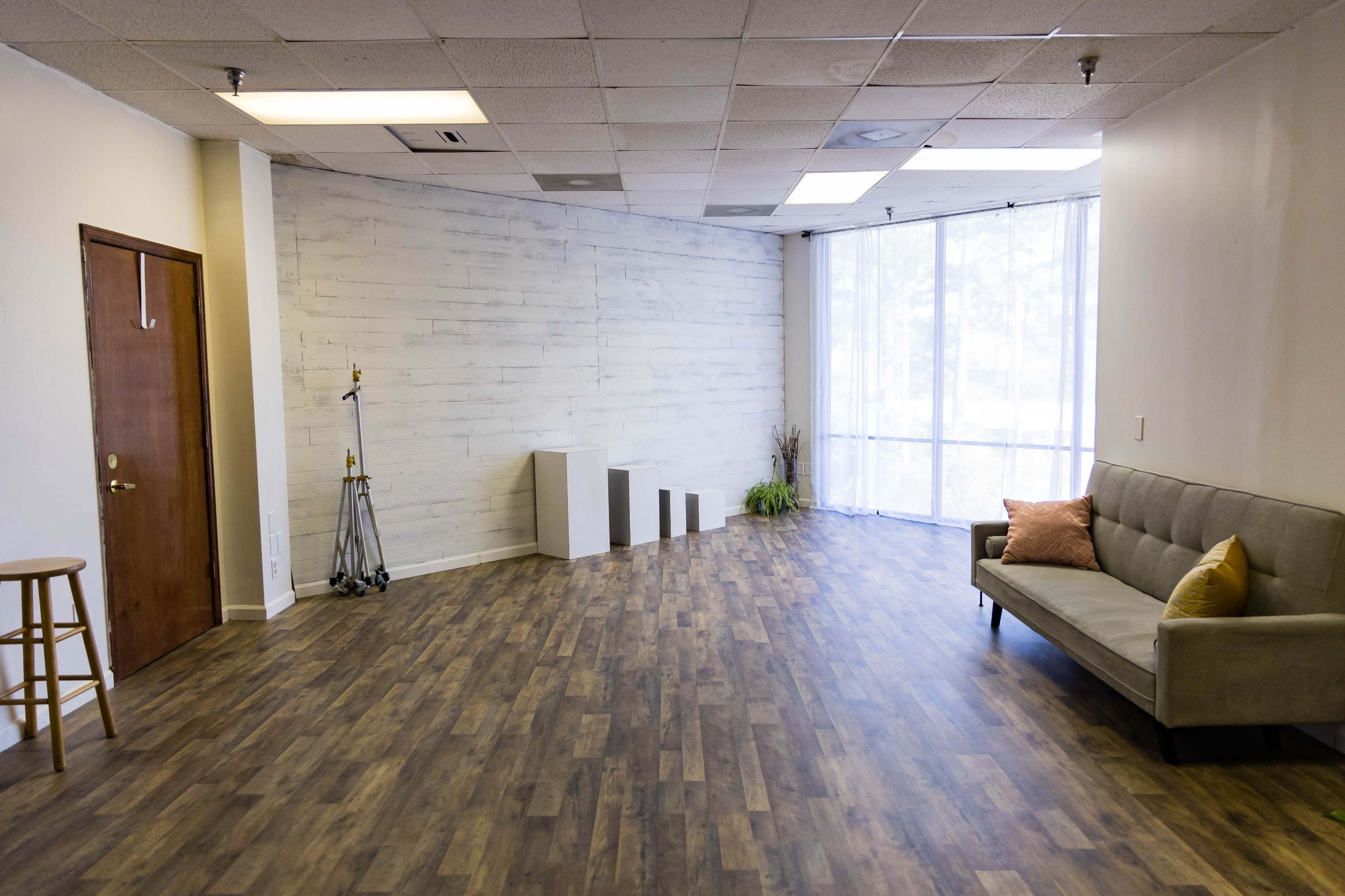 A spacious, empty room with wooden flooring, white walls, a sofa, and natural light coming through large windows.