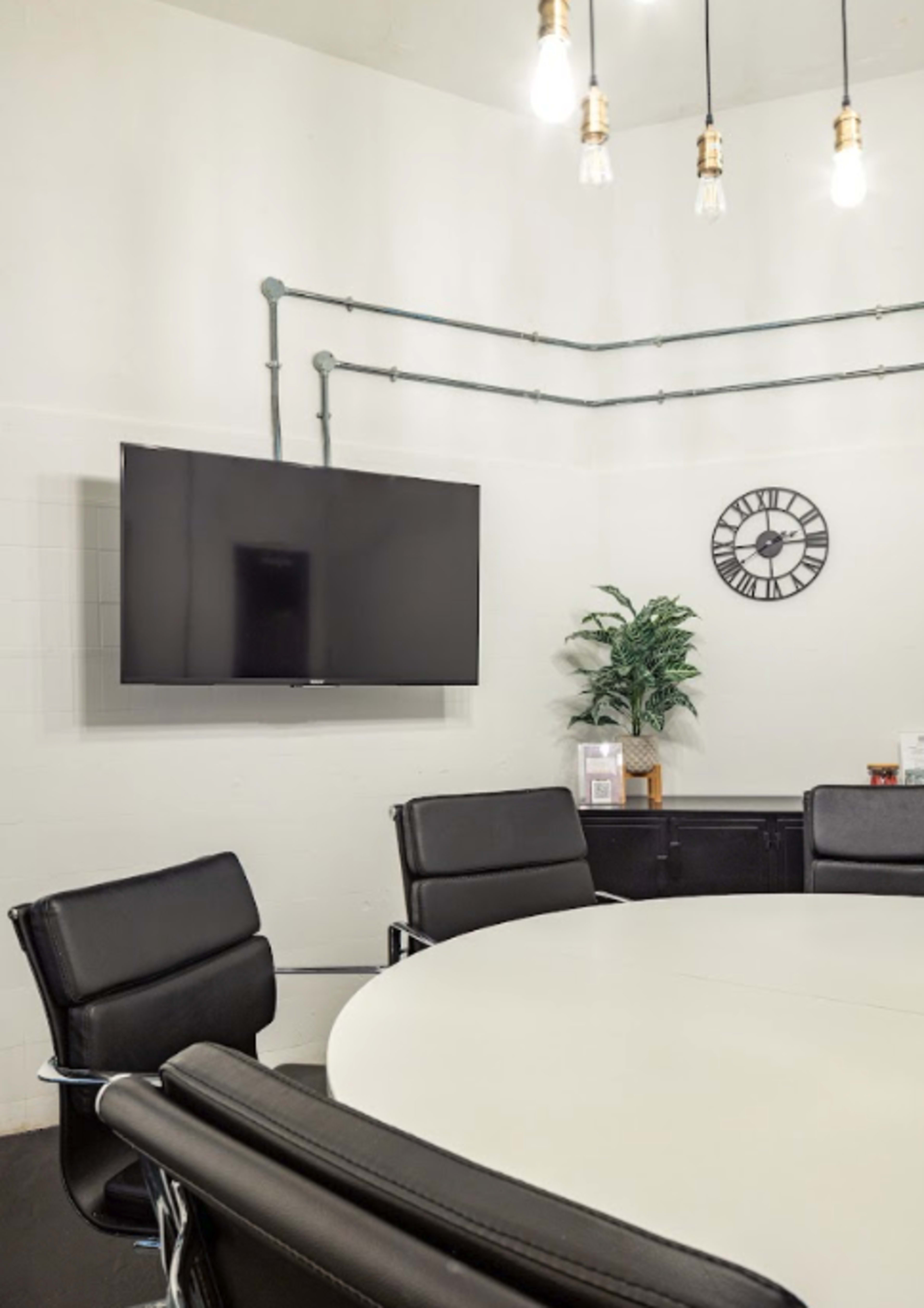 A modern conference room features a circular table, black leather chairs, a wall-mounted TV, and a clock, with industrial pipes and a potted plant.