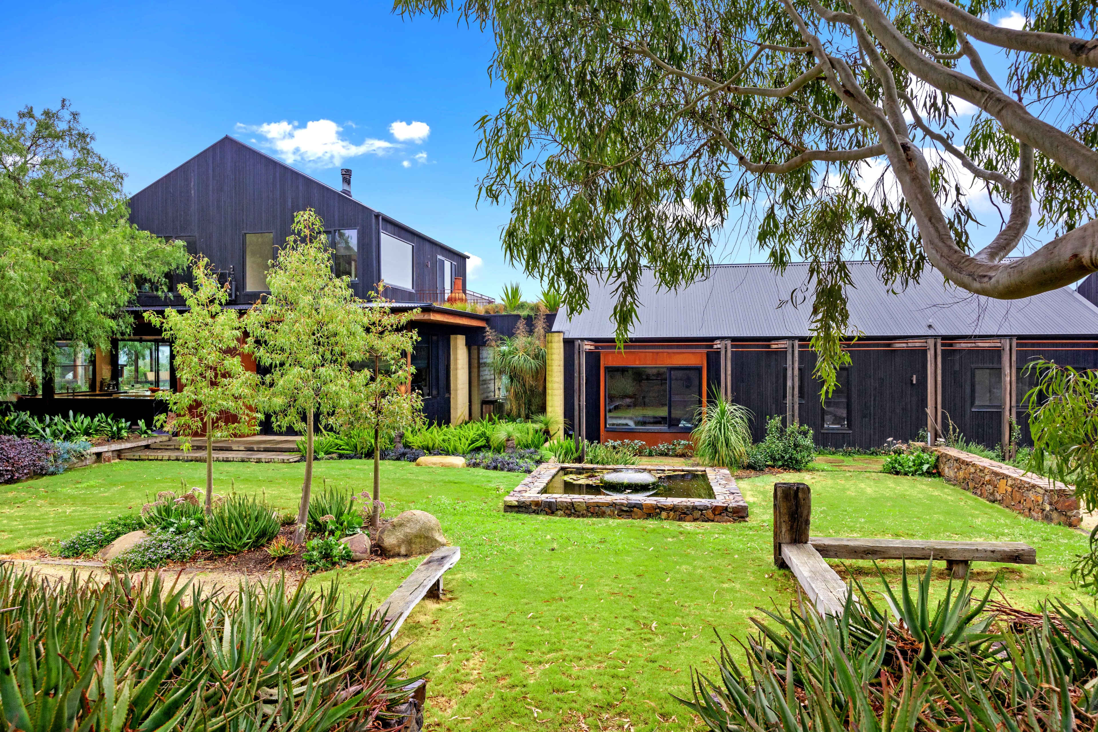 A modern house with a large garden, featuring a pond and landscaped greenery, is surrounded by trees and outdoor seating areas.