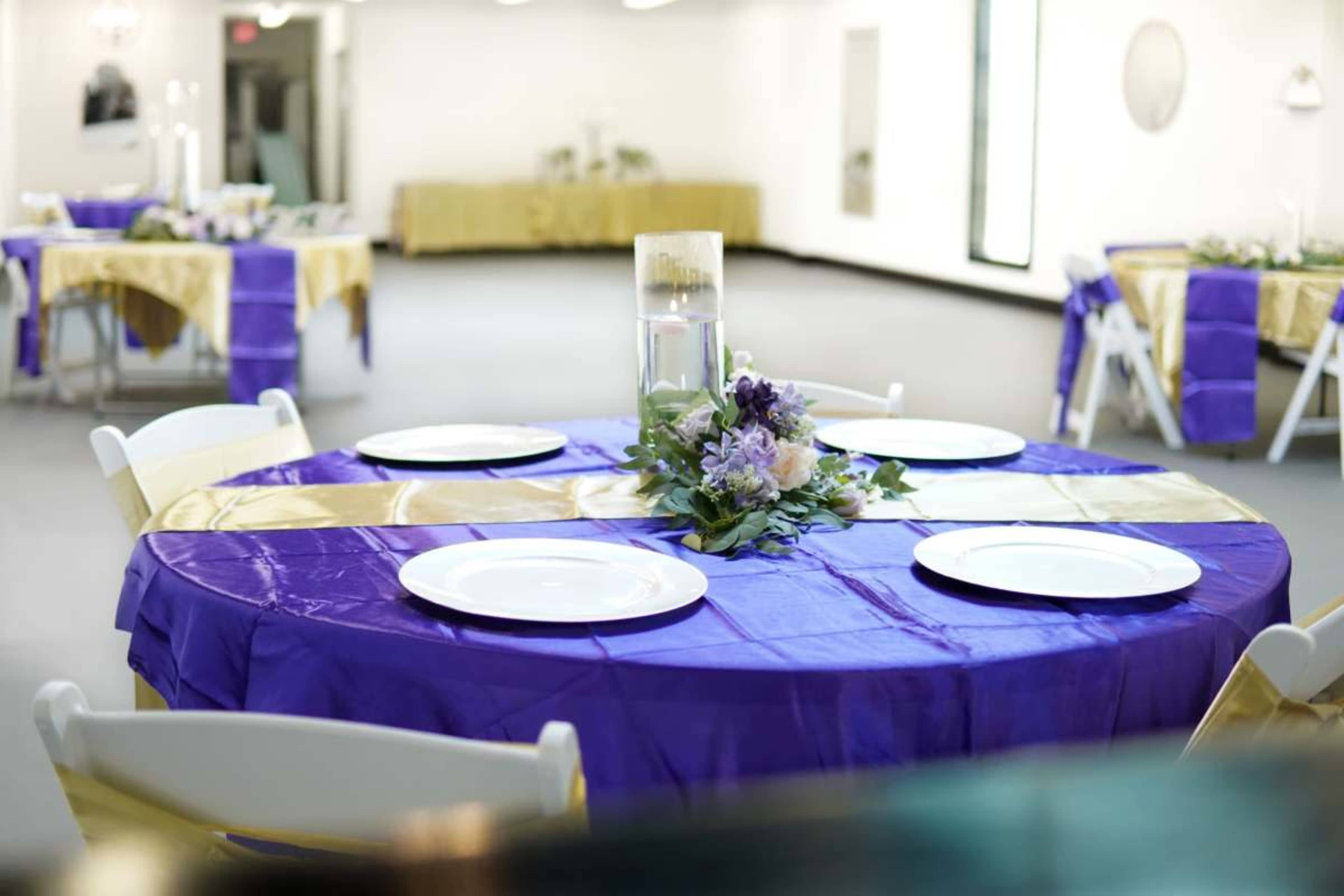 The image shows a banquet hall with circular tables covered in purple and gold tablecloths, each adorned with white plates and a floral centerpiece.