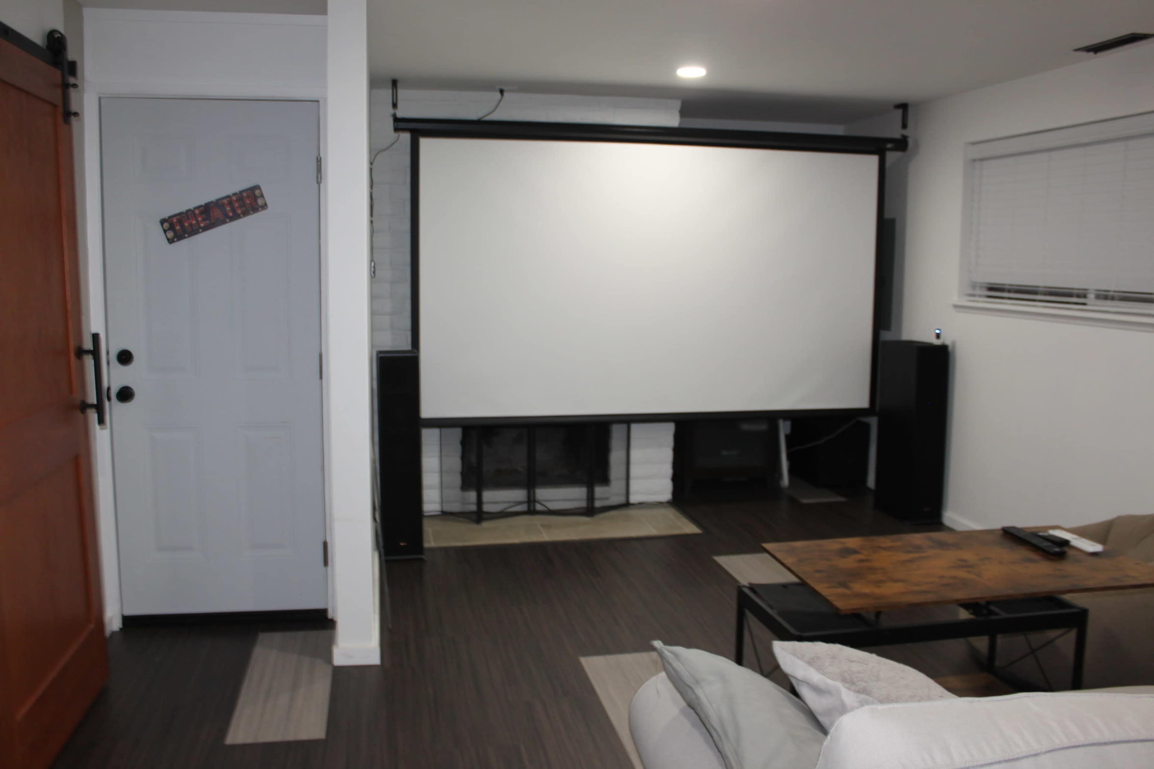 The image shows a room with a large projector screen mounted on the wall, a fireplace, a wooden coffee table, and plush seating.