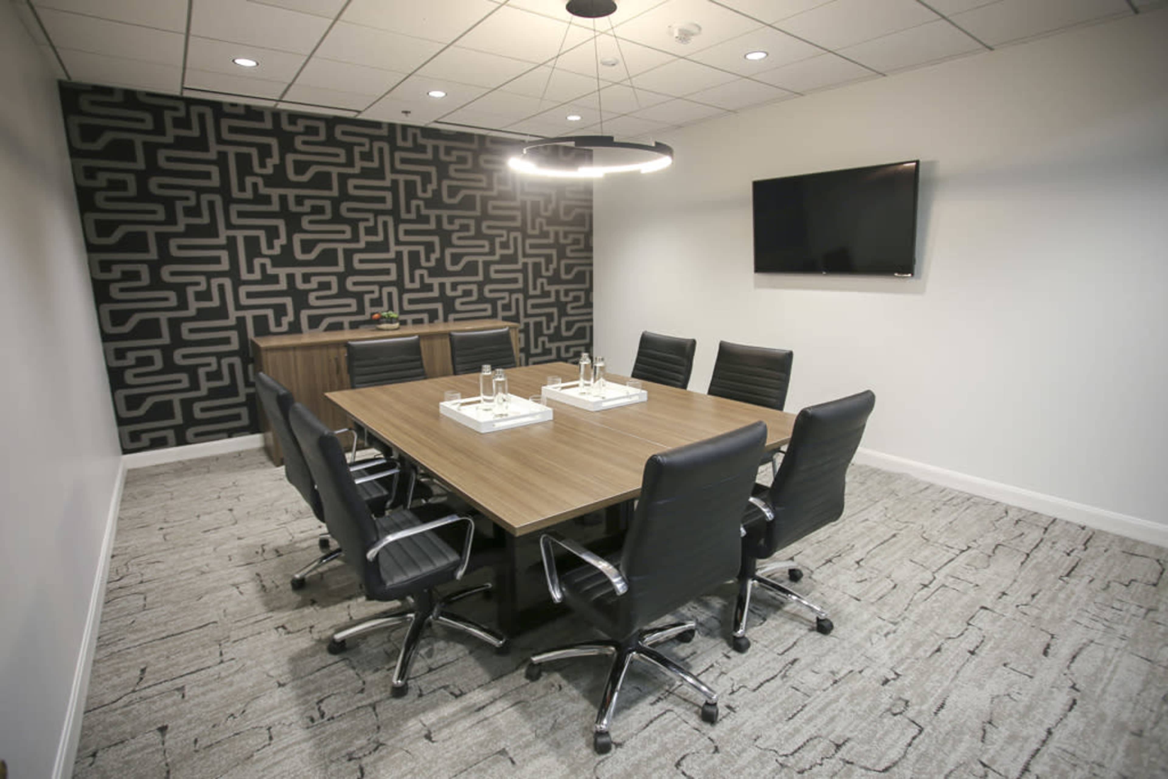 A modern conference room with a large rectangular wooden table surrounded by black office chairs, a patterned accent wall, and a flat-screen TV mounted on the wall.