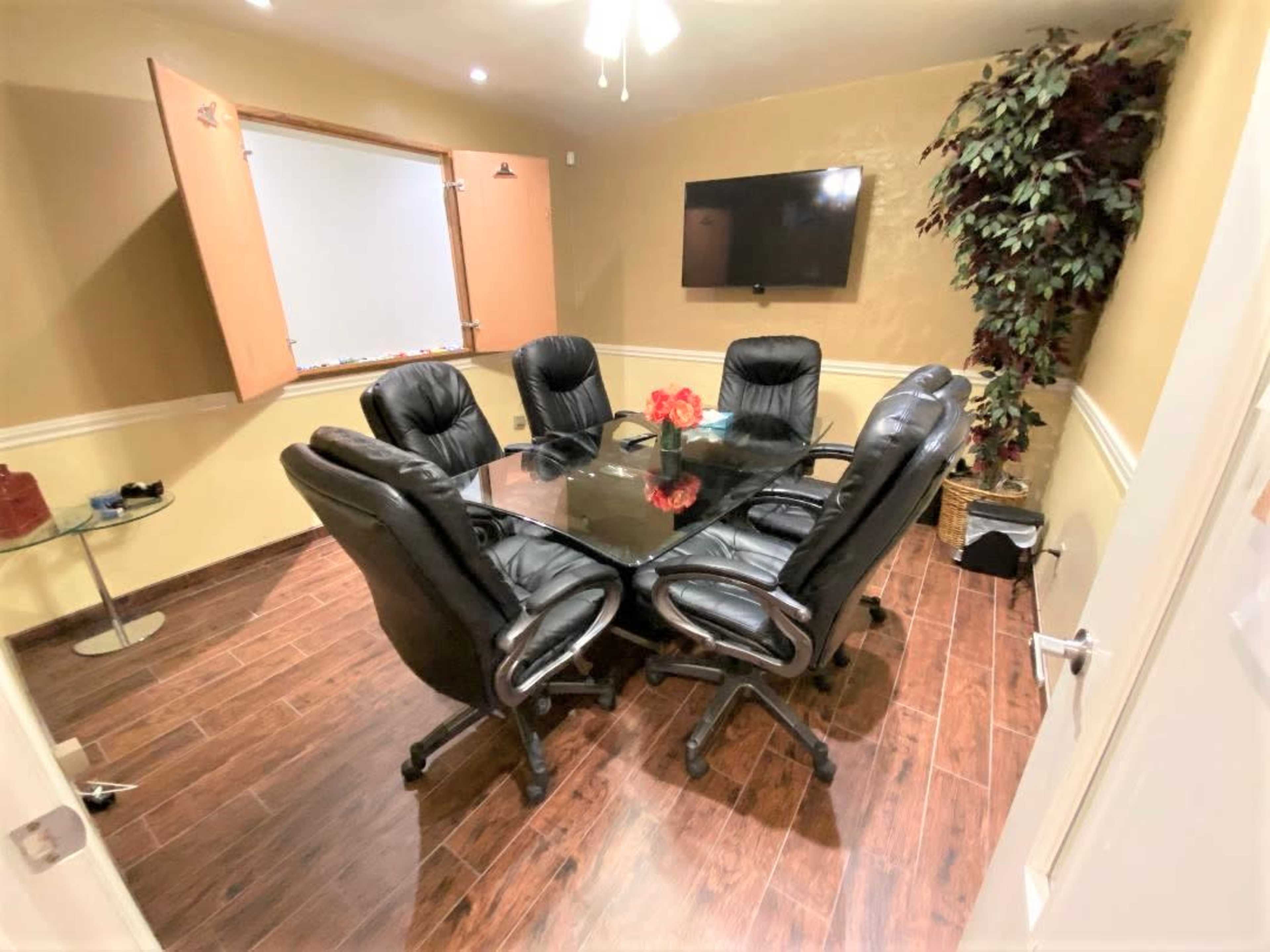 The image shows a small conference room with a round glass table surrounded by six black leather office chairs, a large television on the wall, and a window with shutters.
