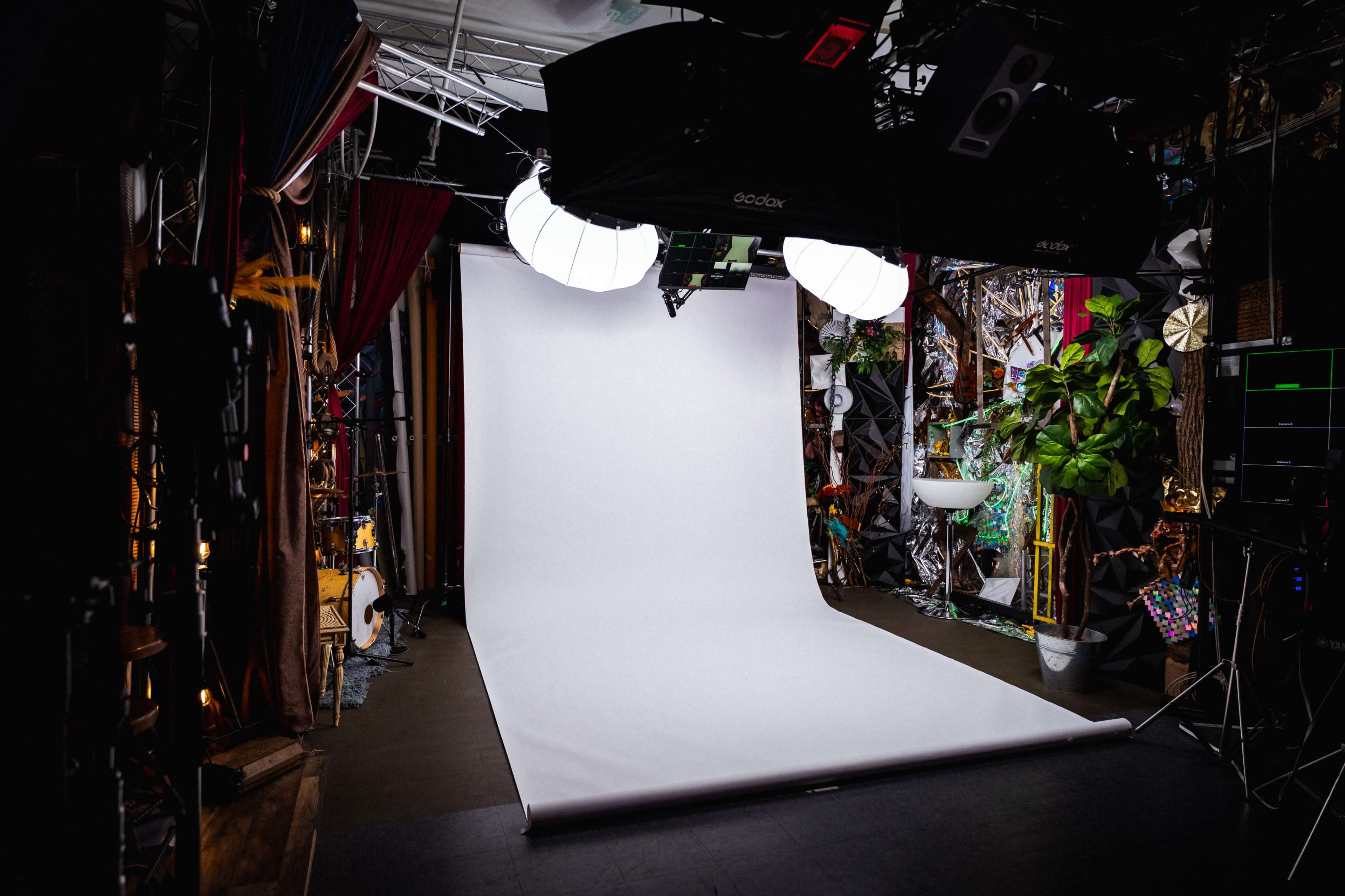 The image shows a photo studio with a white backdrop, large lighting equipment overhead, and various decorative items and plants surrounding the space.