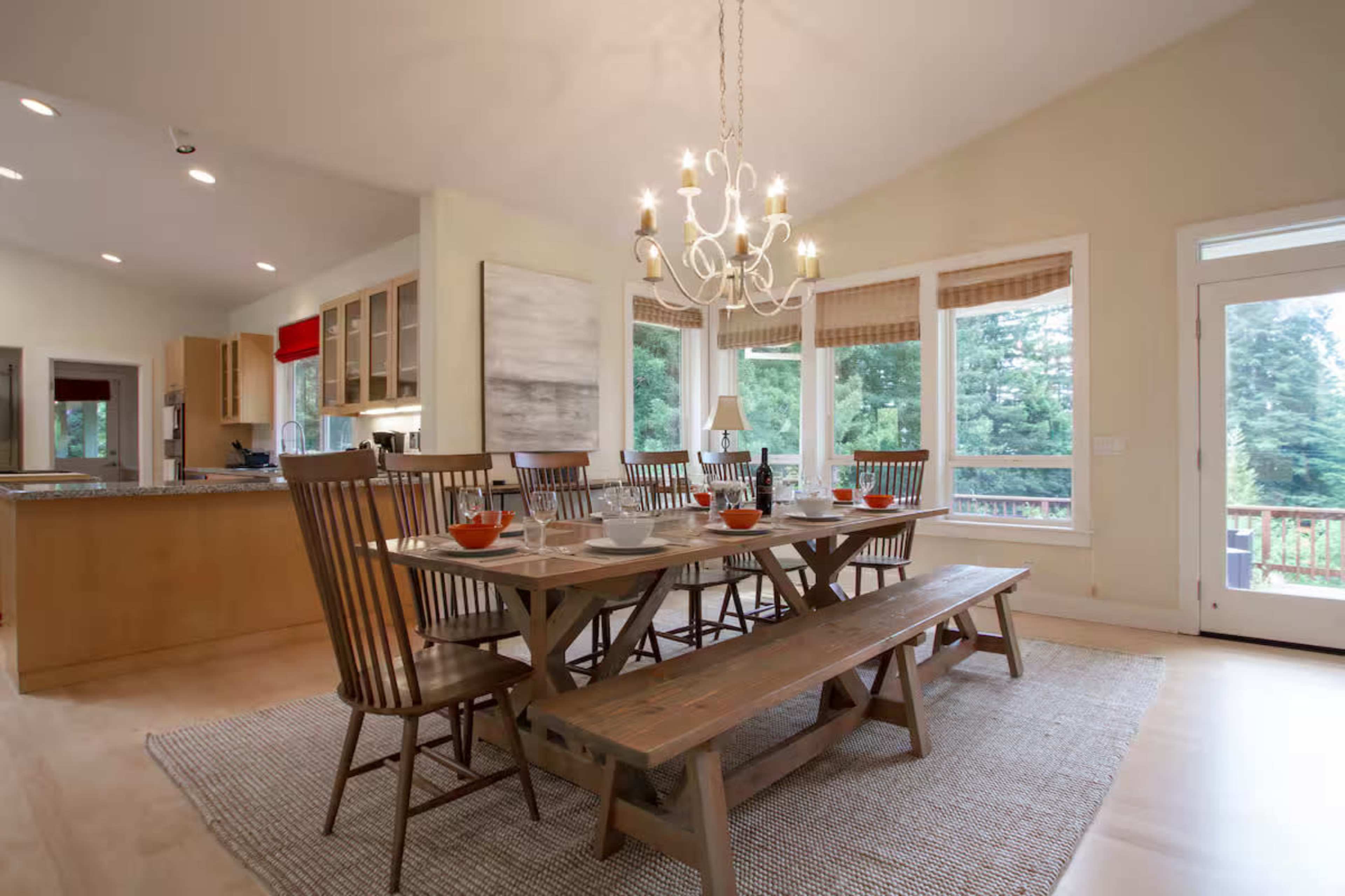 The image shows a spacious dining area with a wooden table set for a meal, surrounded by several chairs, and a large window offering a view of the outdoors.