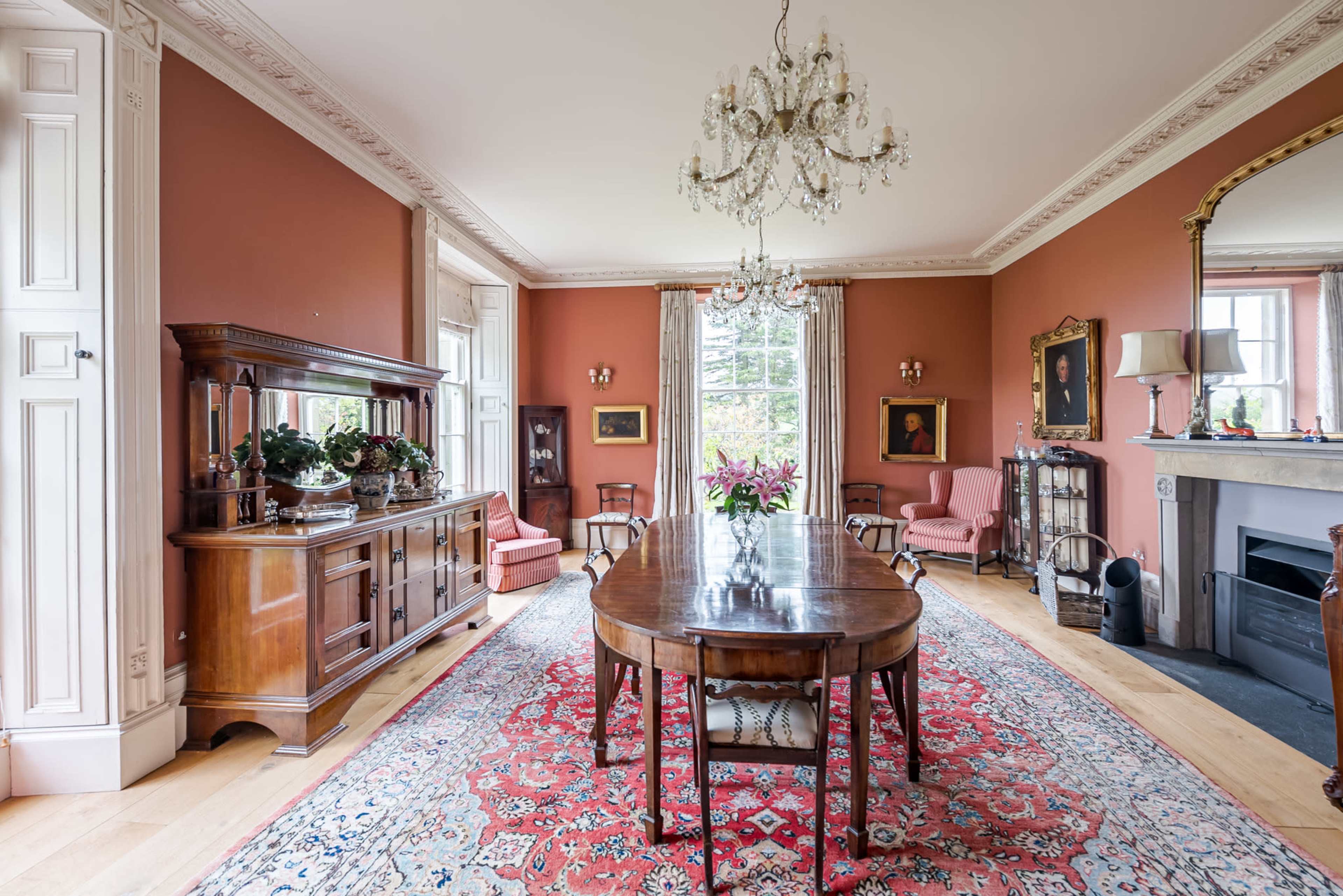 The image shows a formal dining room with a wooden table, chairs, a chandelier, and a patterned rug, complemented by artwork and a buffet cabinet.