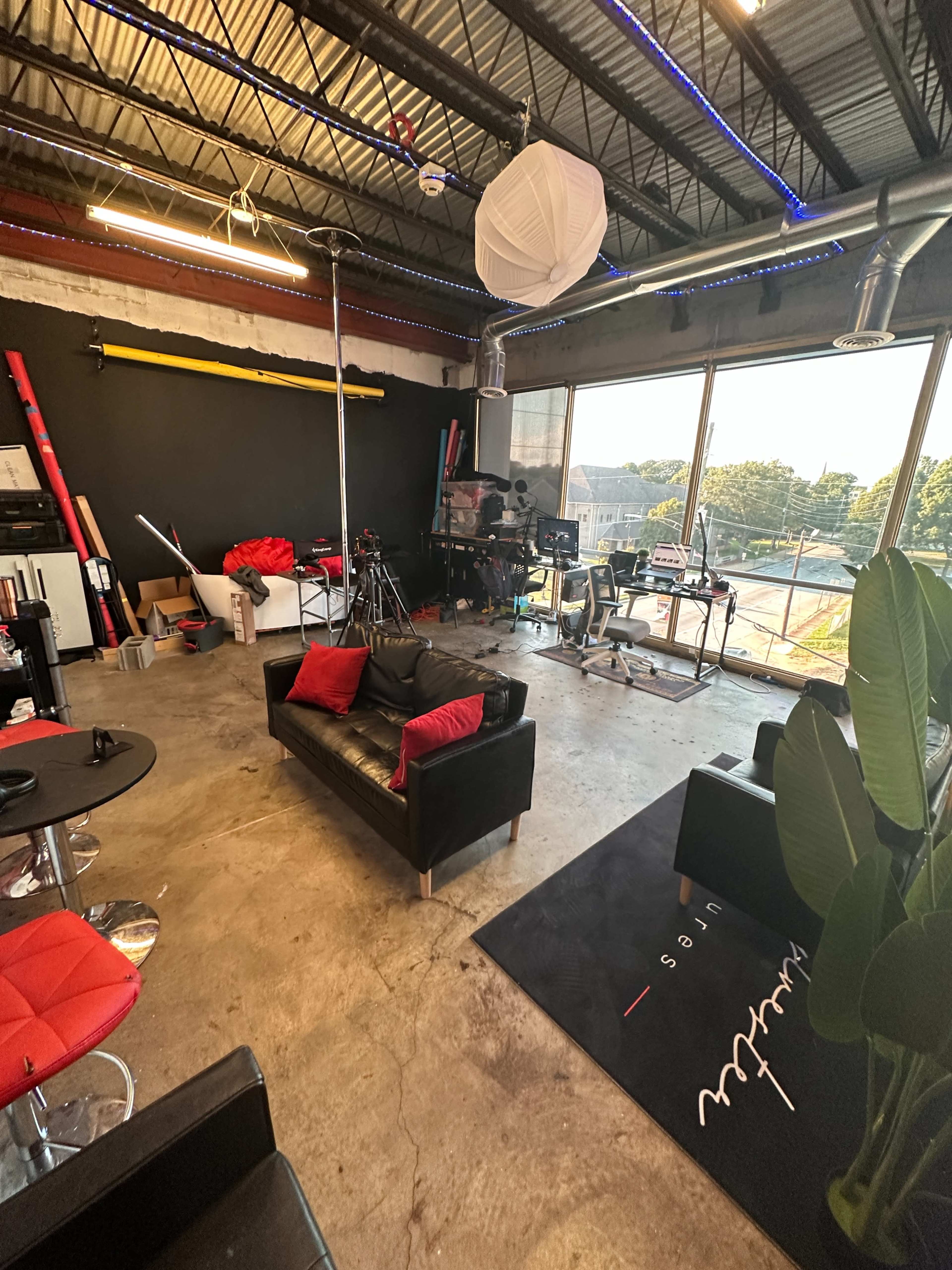 A studio space featuring black leather sofas, a workspace with cameras and equipment, and large windows letting in natural light.