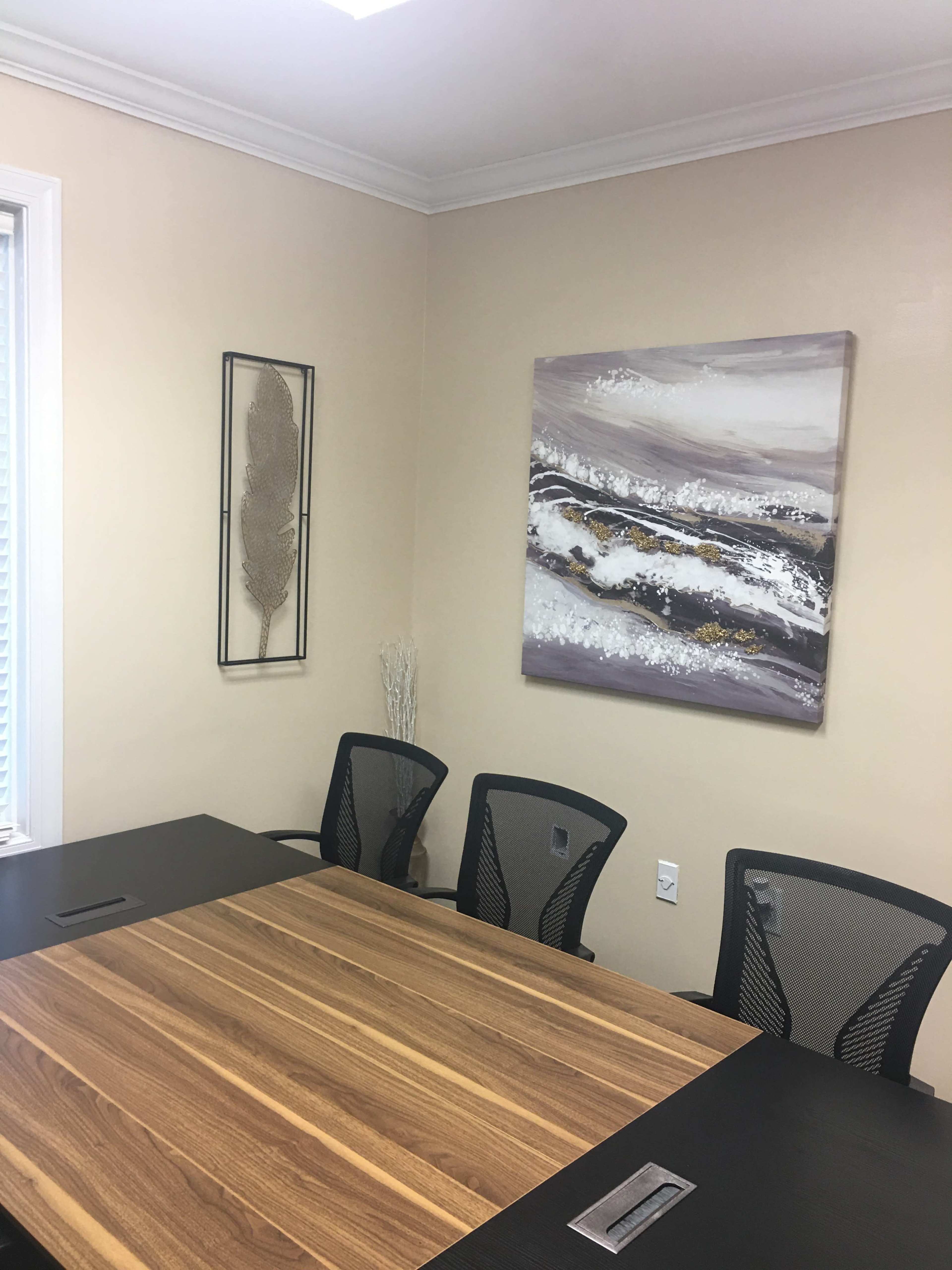 A conference room with a large wooden table and four black mesh chairs, accompanied by a wall decoration and a painting.
