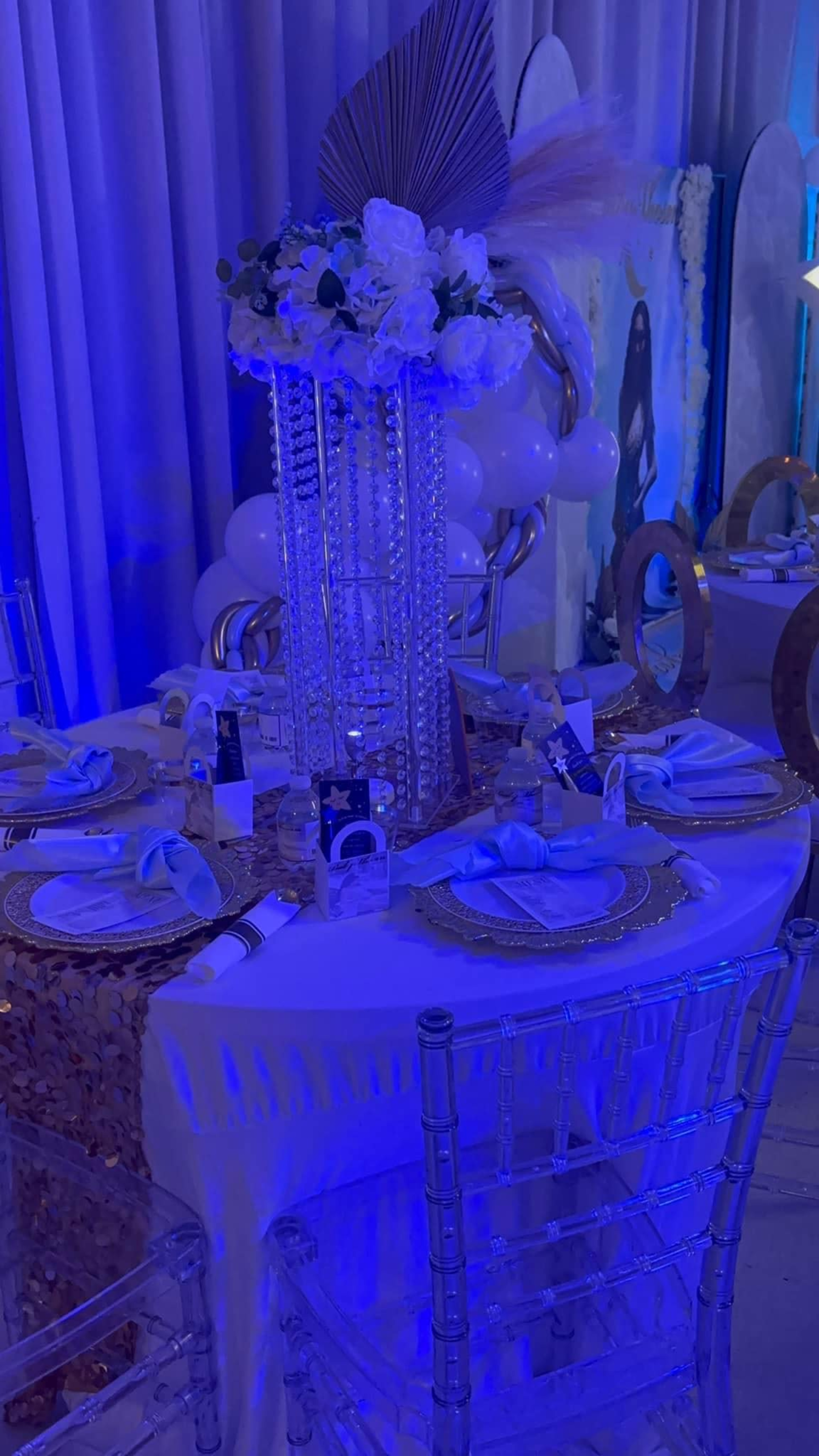 A elegantly set table with a tall floral centerpiece, surrounded by clear chiavari chairs and decorated with blue lighting.