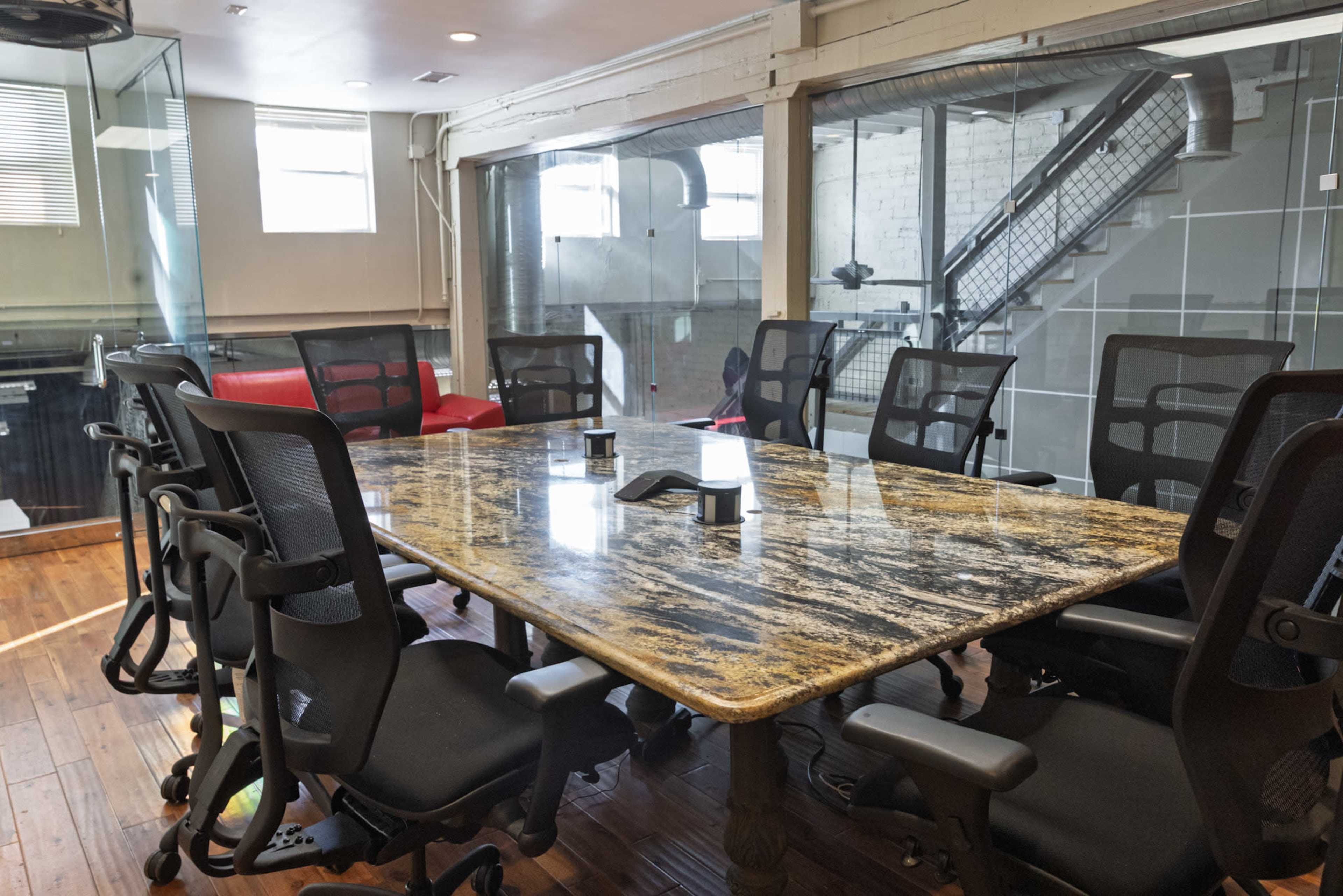 A modern conference room features a large granite table surrounded by ergonomic black chairs, with glass walls and a staircase visible in the background.