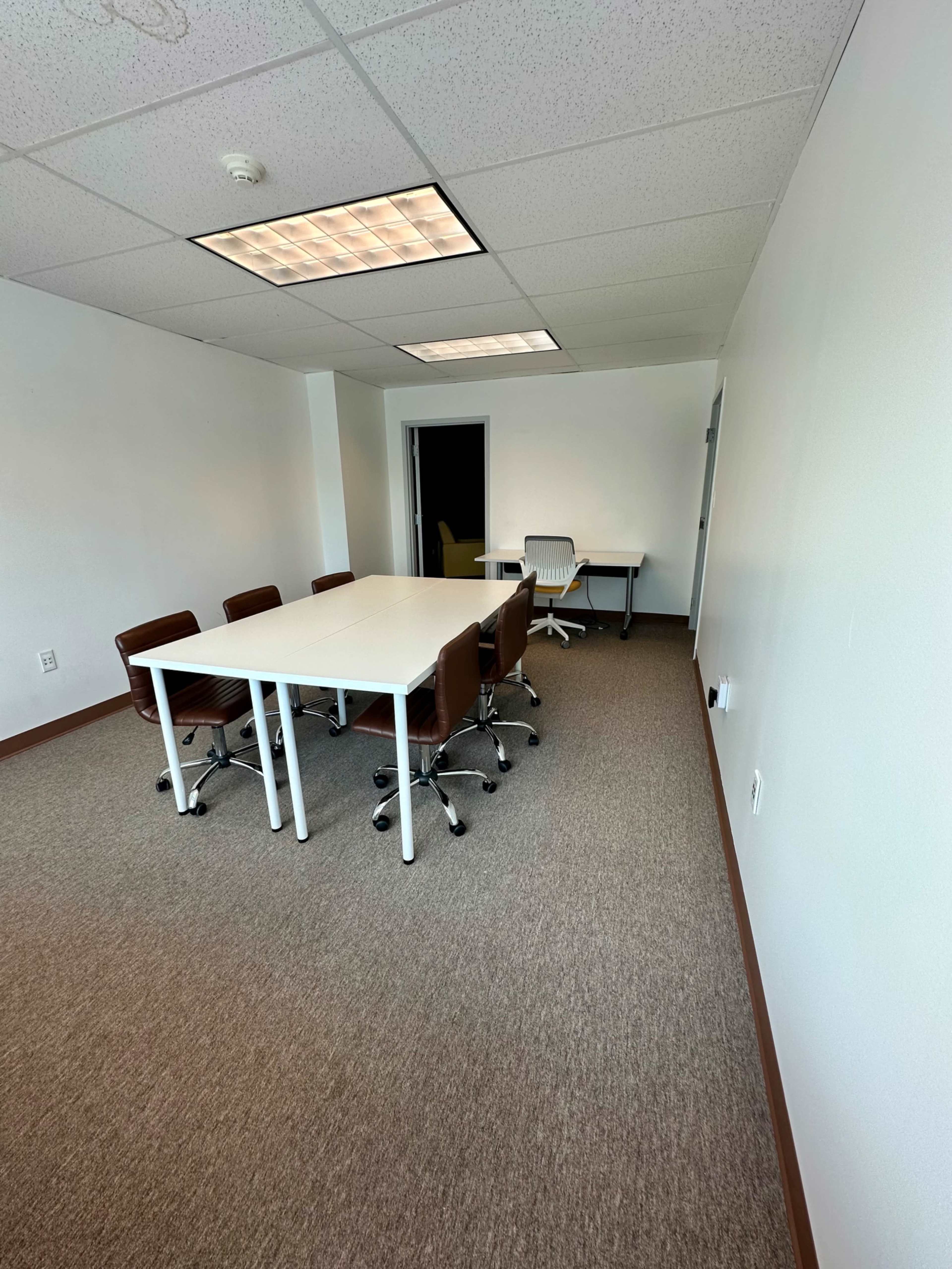 A conference room with a white table surrounded by brown chairs and an additional desk in the corner.