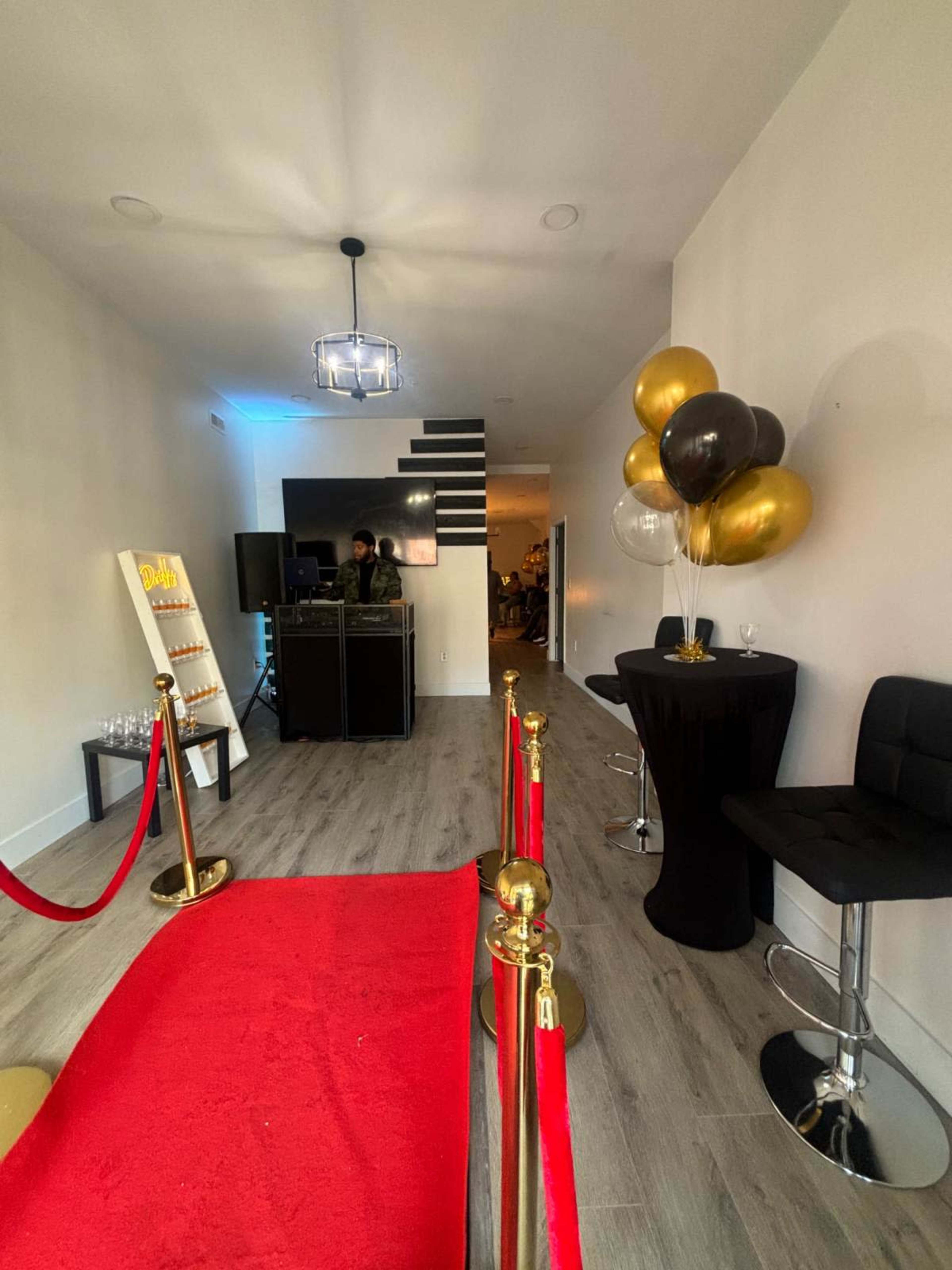 The image shows a festive event space featuring a red carpet entrance, a DJ setup, a table with balloons, and a lounge area.
