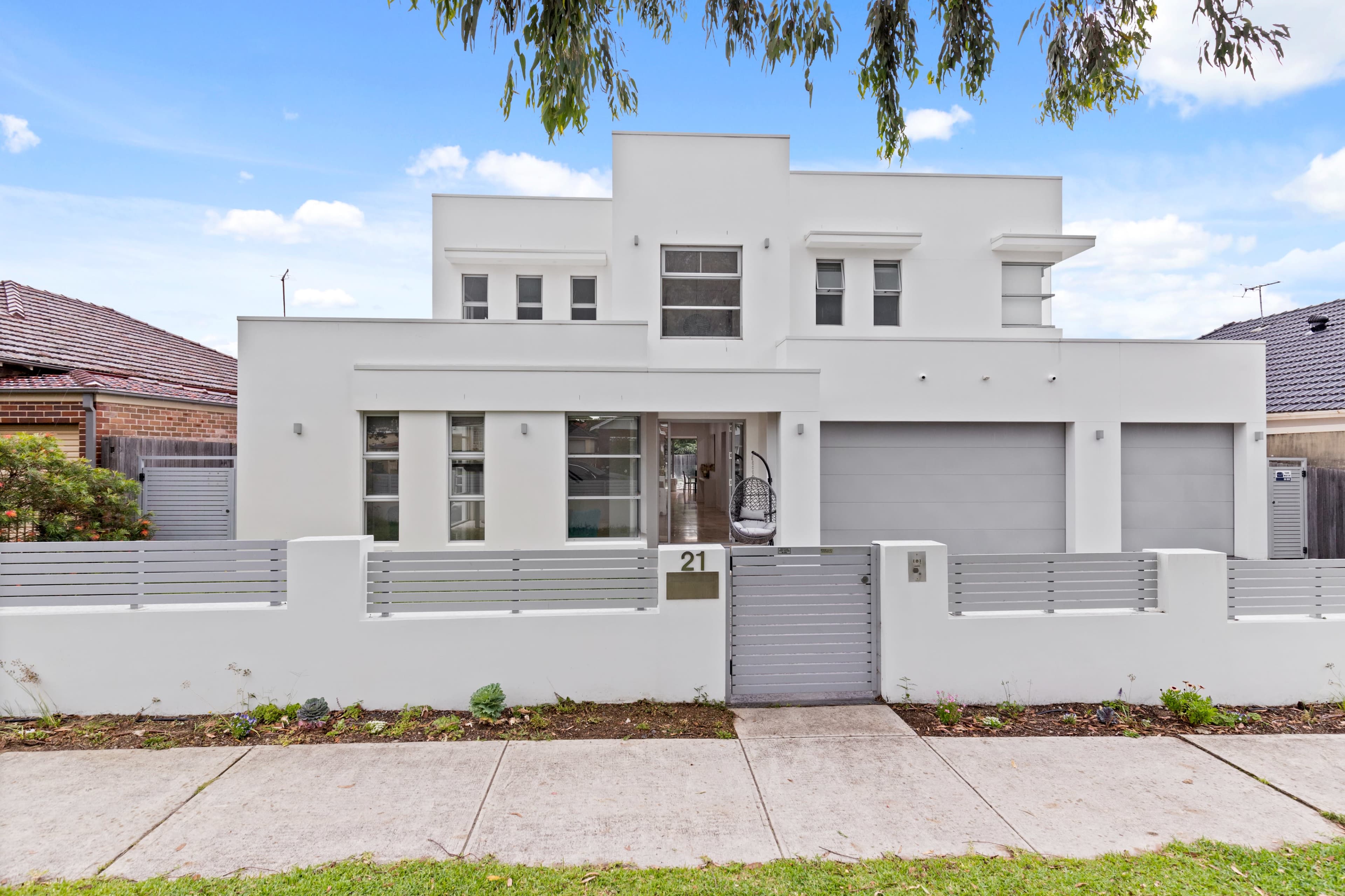 The image shows a modern two-story white house with a front gate and driveway, situated on a sidewalk lined with grass.
