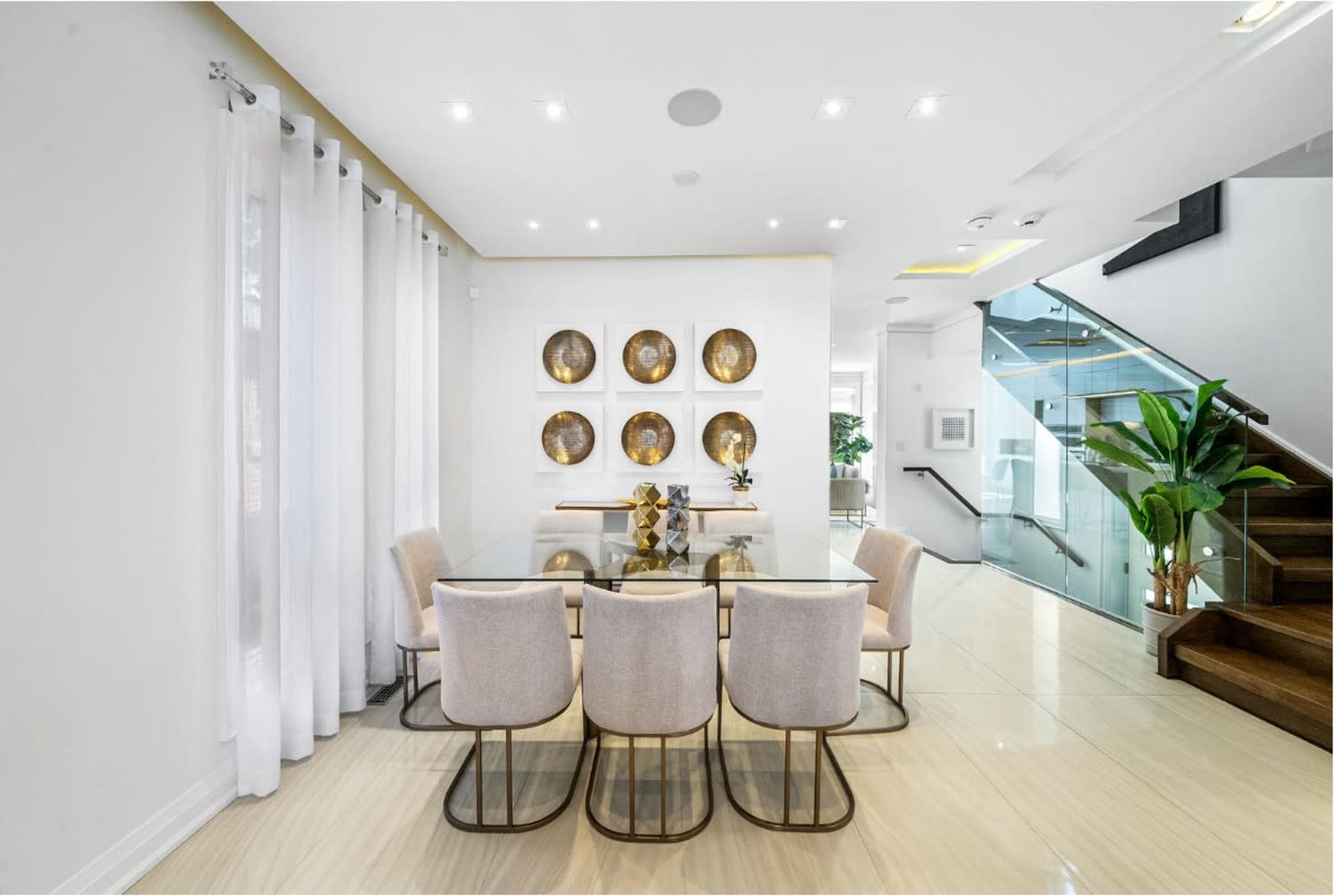 A modern dining area features a glass table surrounded by six upholstered chairs, with decorative wall plates and a staircase in the background.