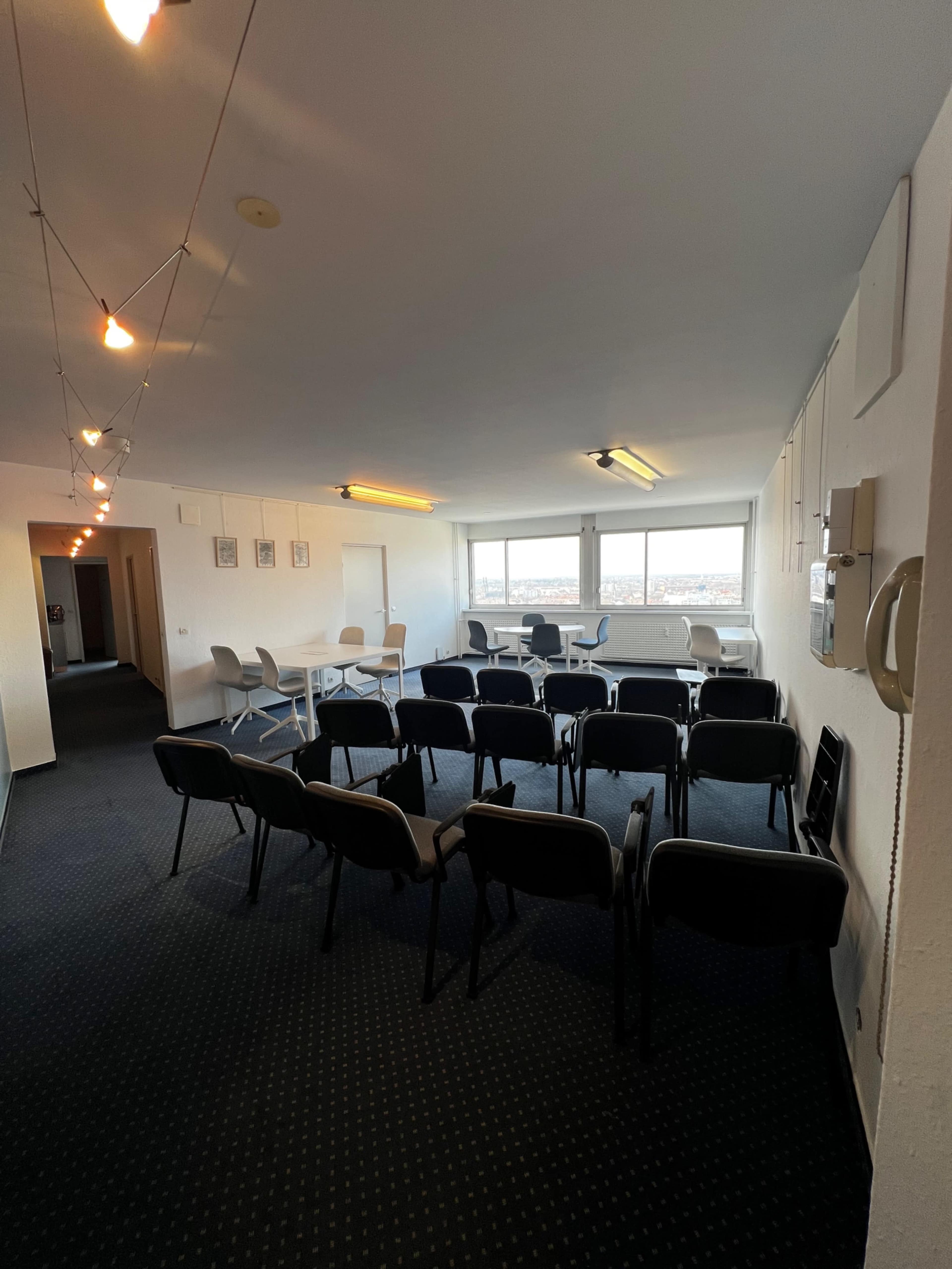 The image shows a spacious room arranged with rows of chairs facing a set of tables and large windows, illuminated by string lights.