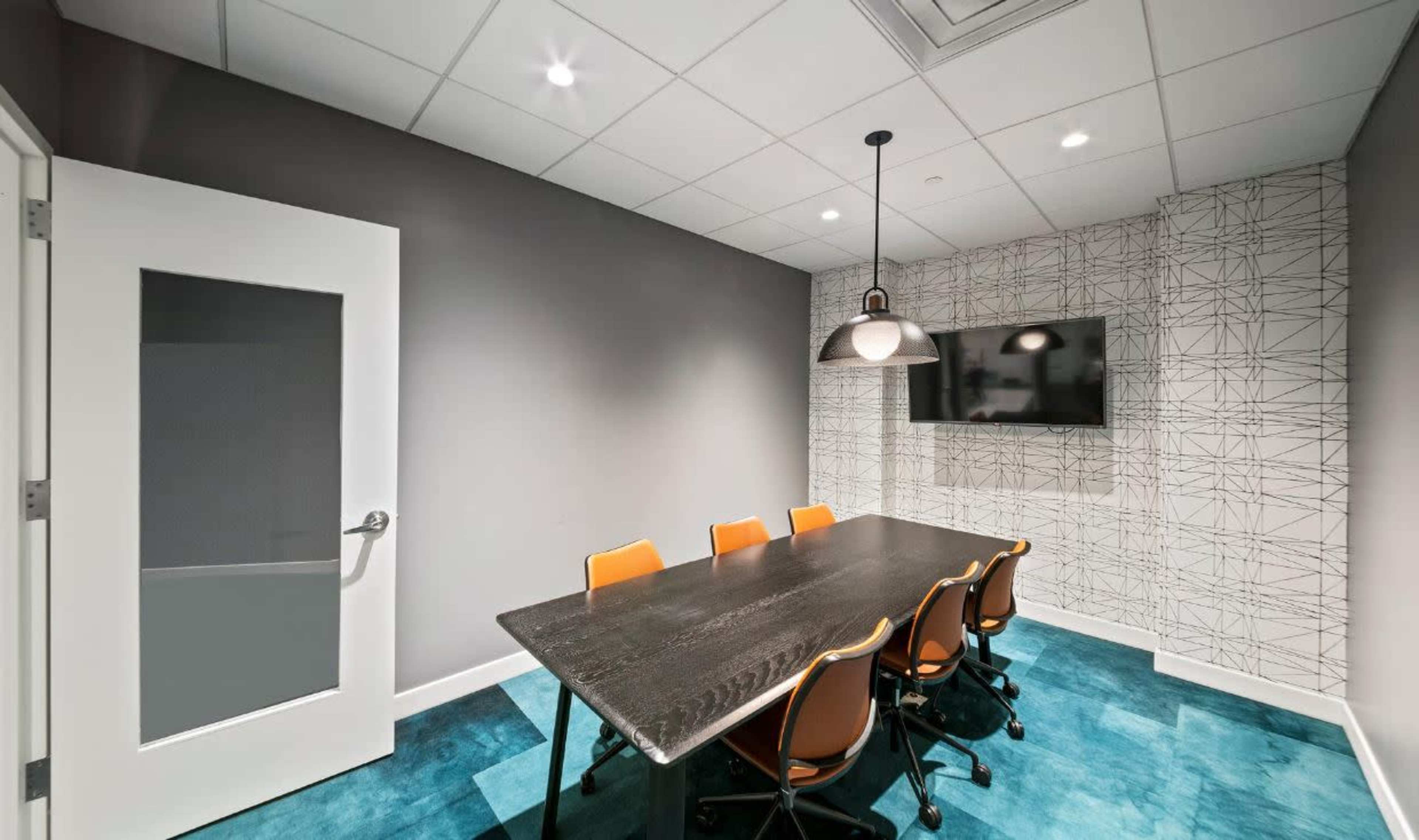 A small conference room features a long black table surrounded by orange chairs, a wall-mounted TV, and patterned wallpaper with a geometric design.