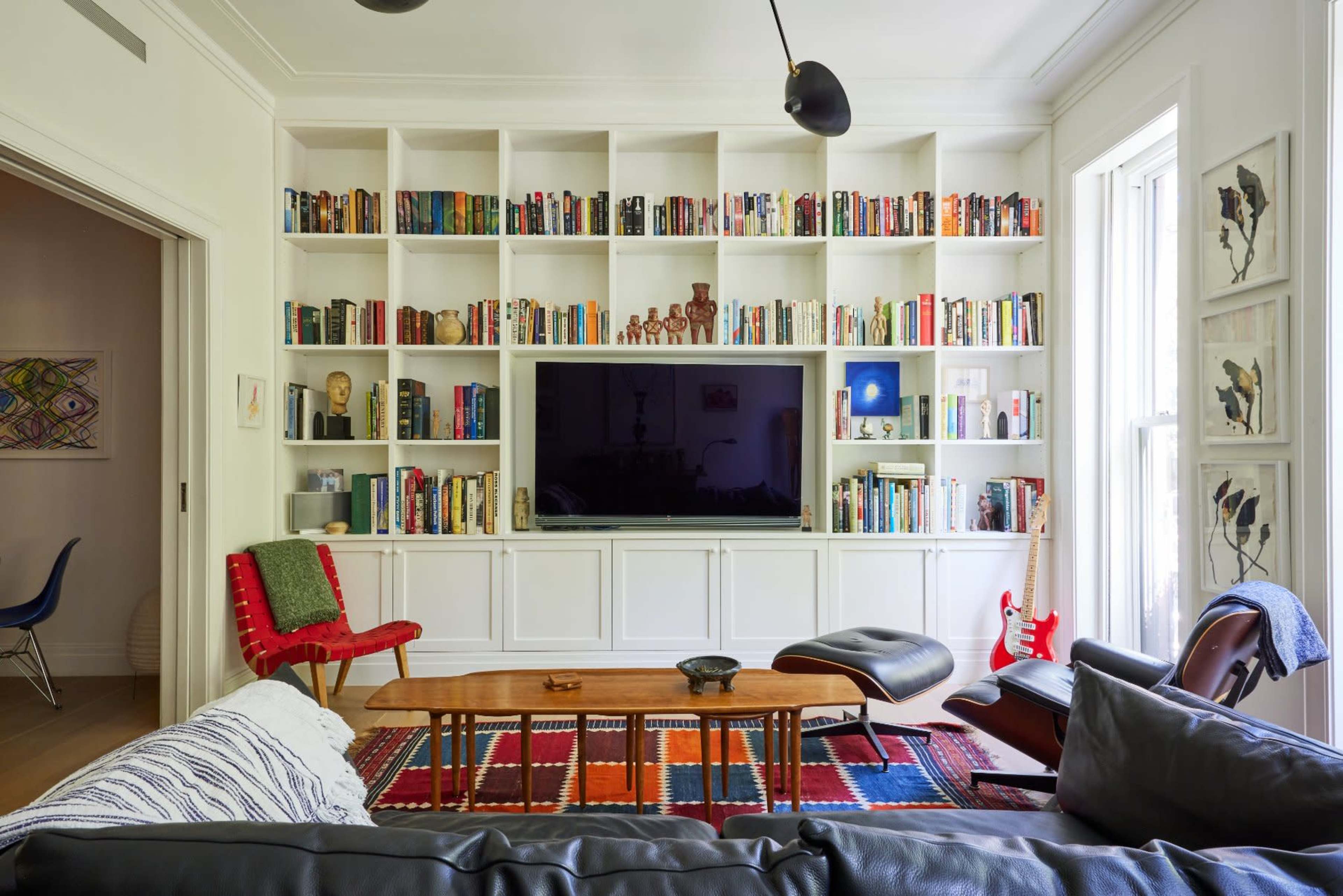 A modern living room features a large bookshelf filled with books, a flat-screen TV, and various seating options arranged around a central coffee table on a colorful rug.