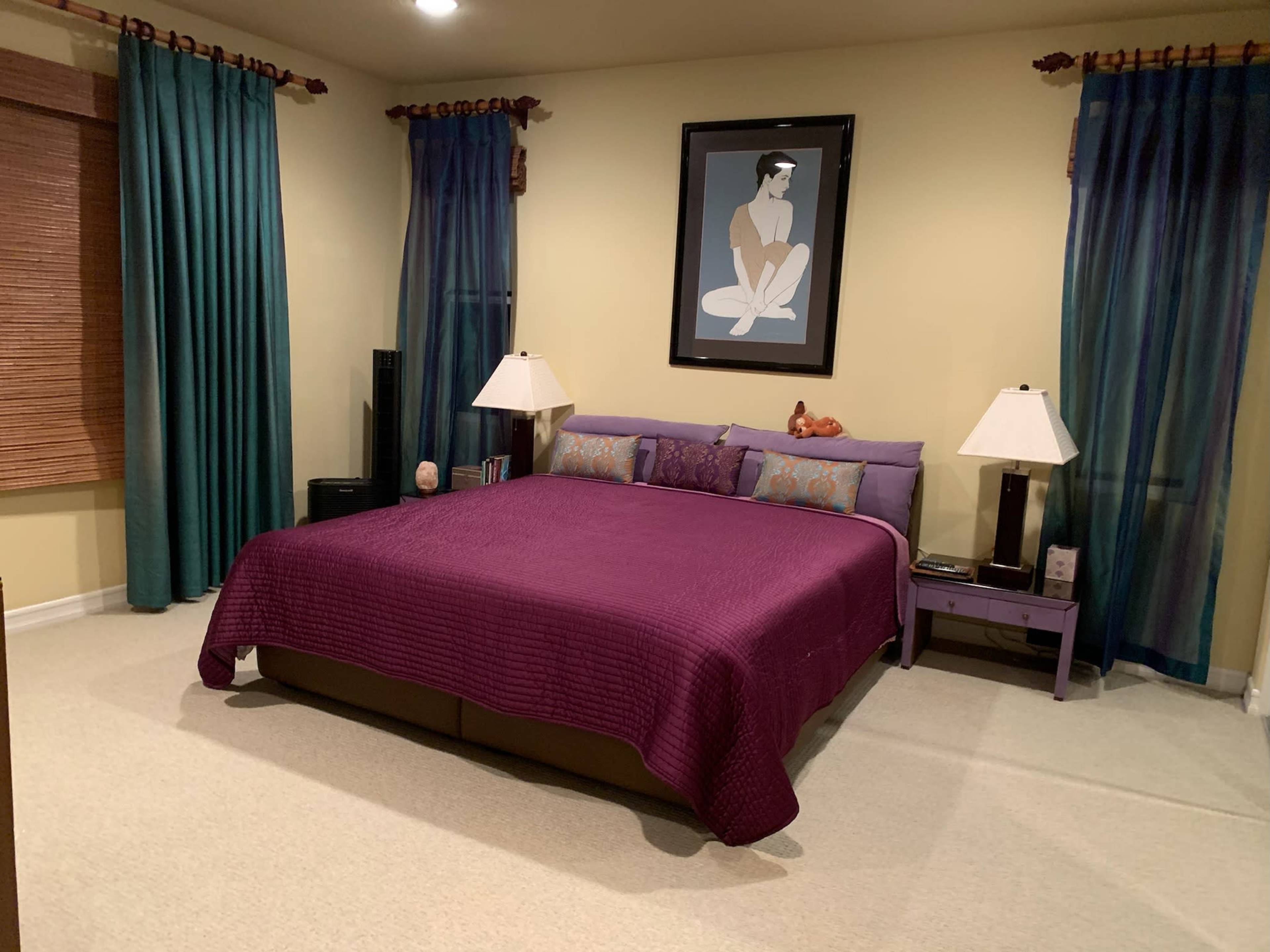 The image shows a neatly arranged bedroom featuring a large purple bedspread on a bed, two bedside lamps, and curtains on the windows.