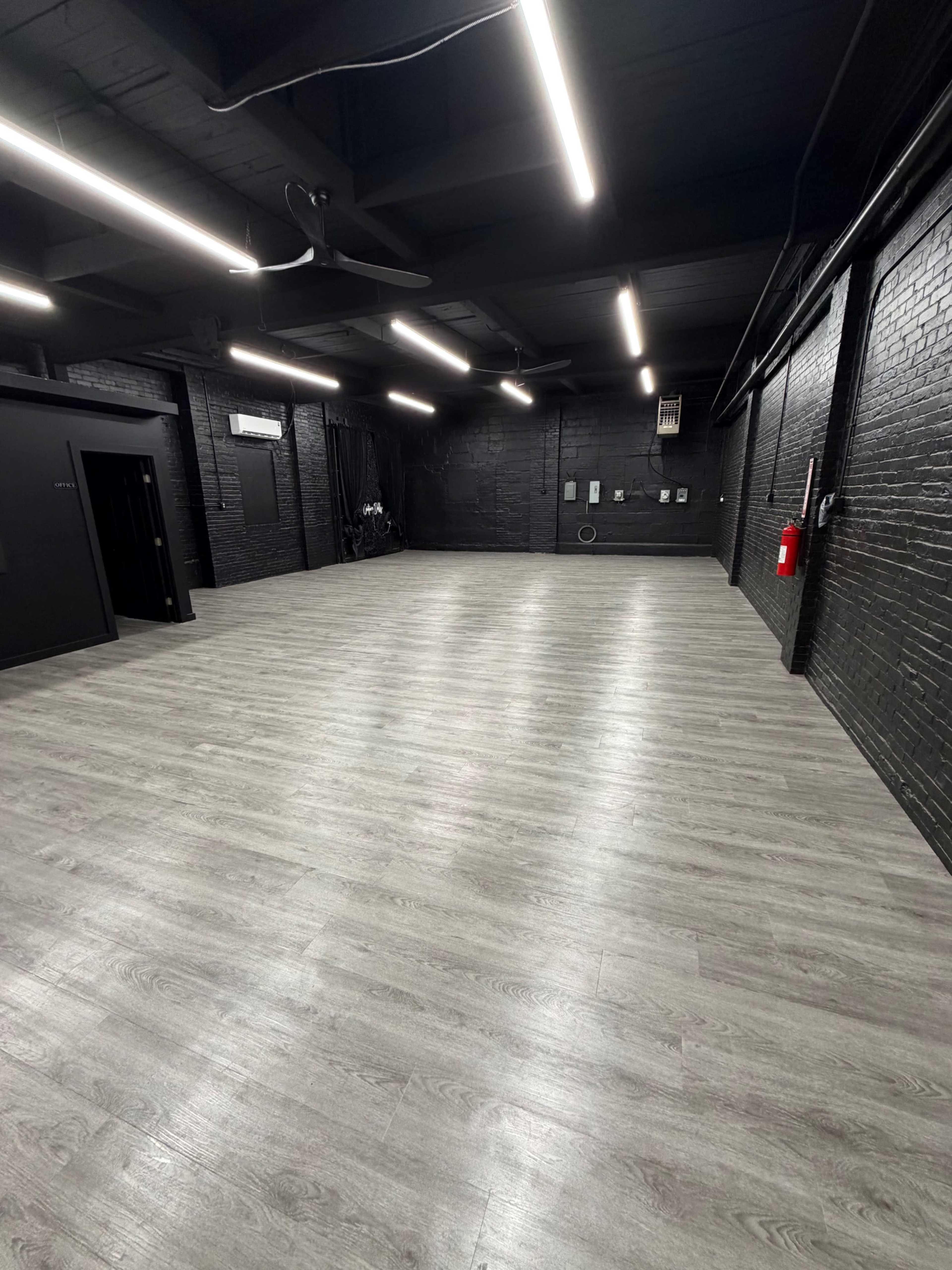 The image shows a spacious, empty room with dark brick walls and grey wooden flooring, illuminated by bright overhead lights.