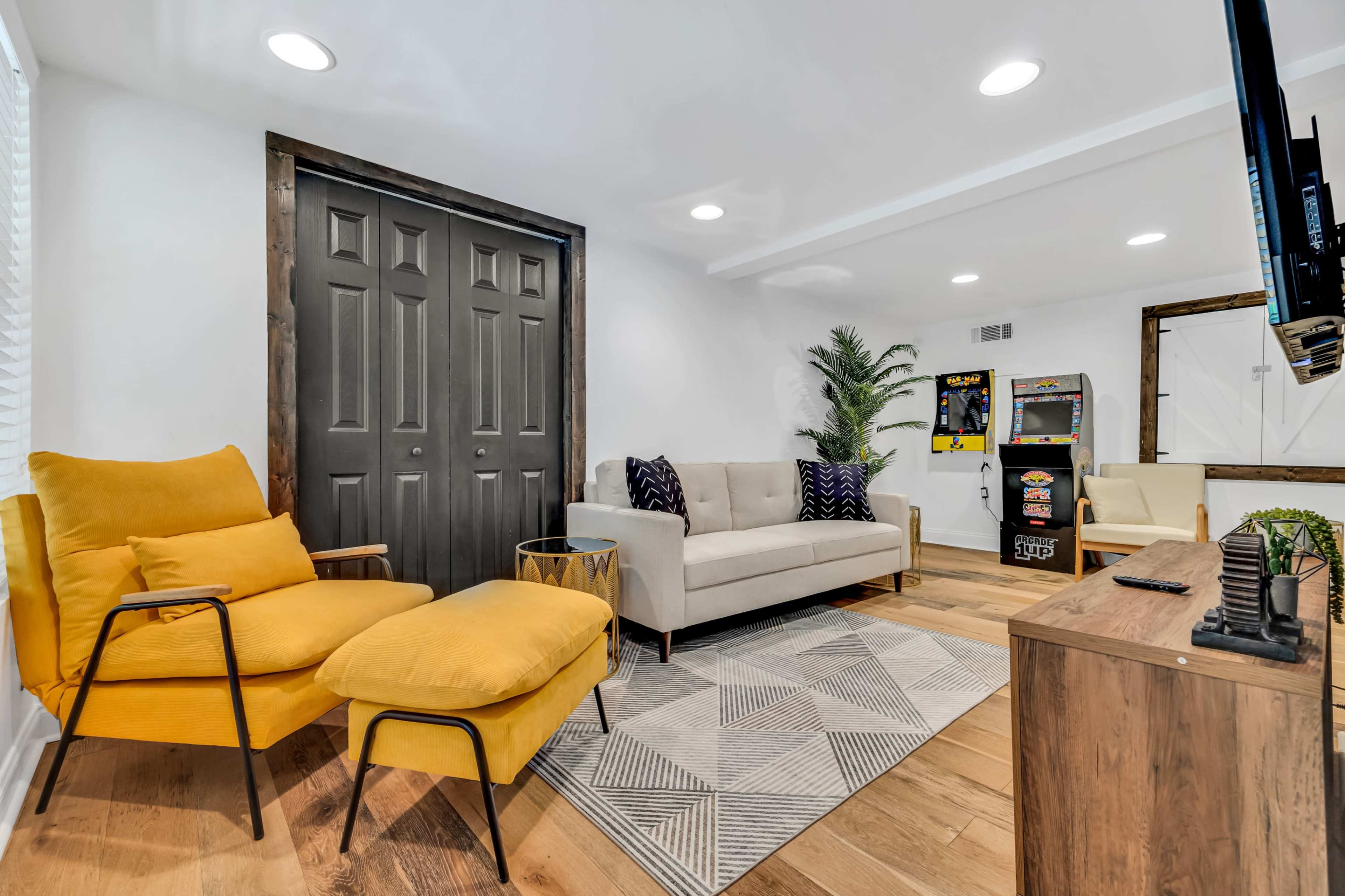 A cozy living room with a yellow armchair, a beige sofa, a patterned rug, and an arcade game machine in the corner.