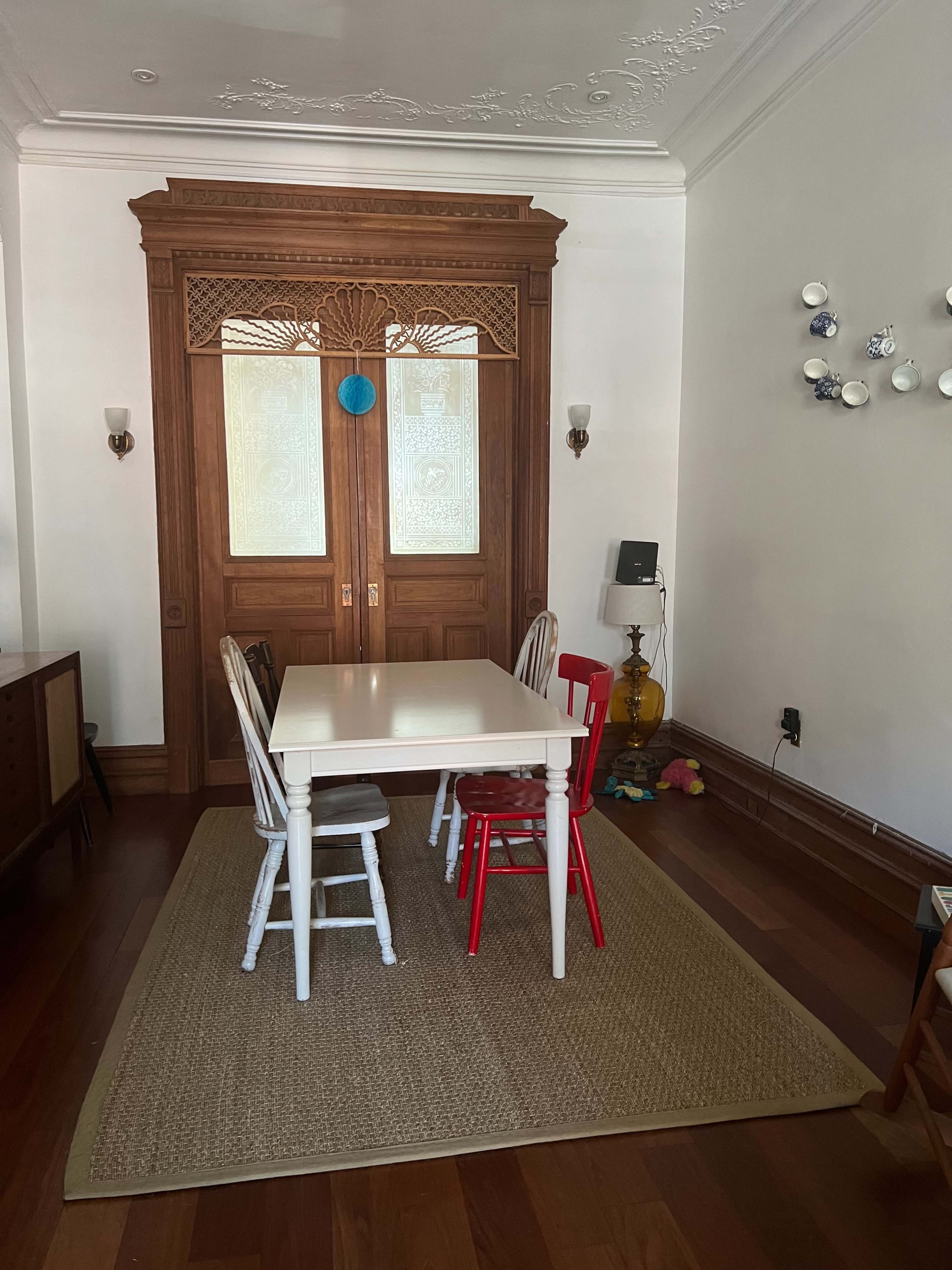 A simple dining area features a white table surrounded by four chairs, with a wooden door and decorative wall elements in the background.