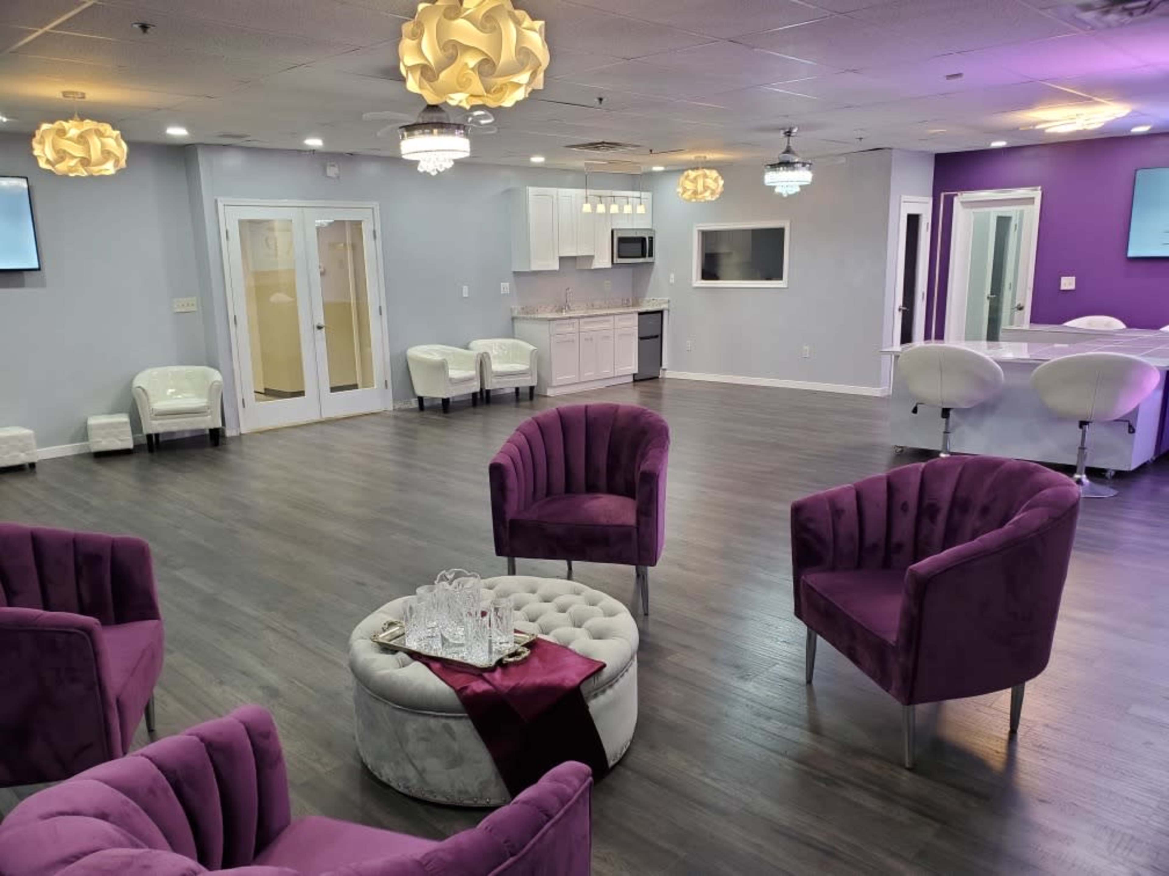 A stylish room with purple accent chairs, a circular ottoman, and a kitchenette, illuminated by decorative light fixtures.