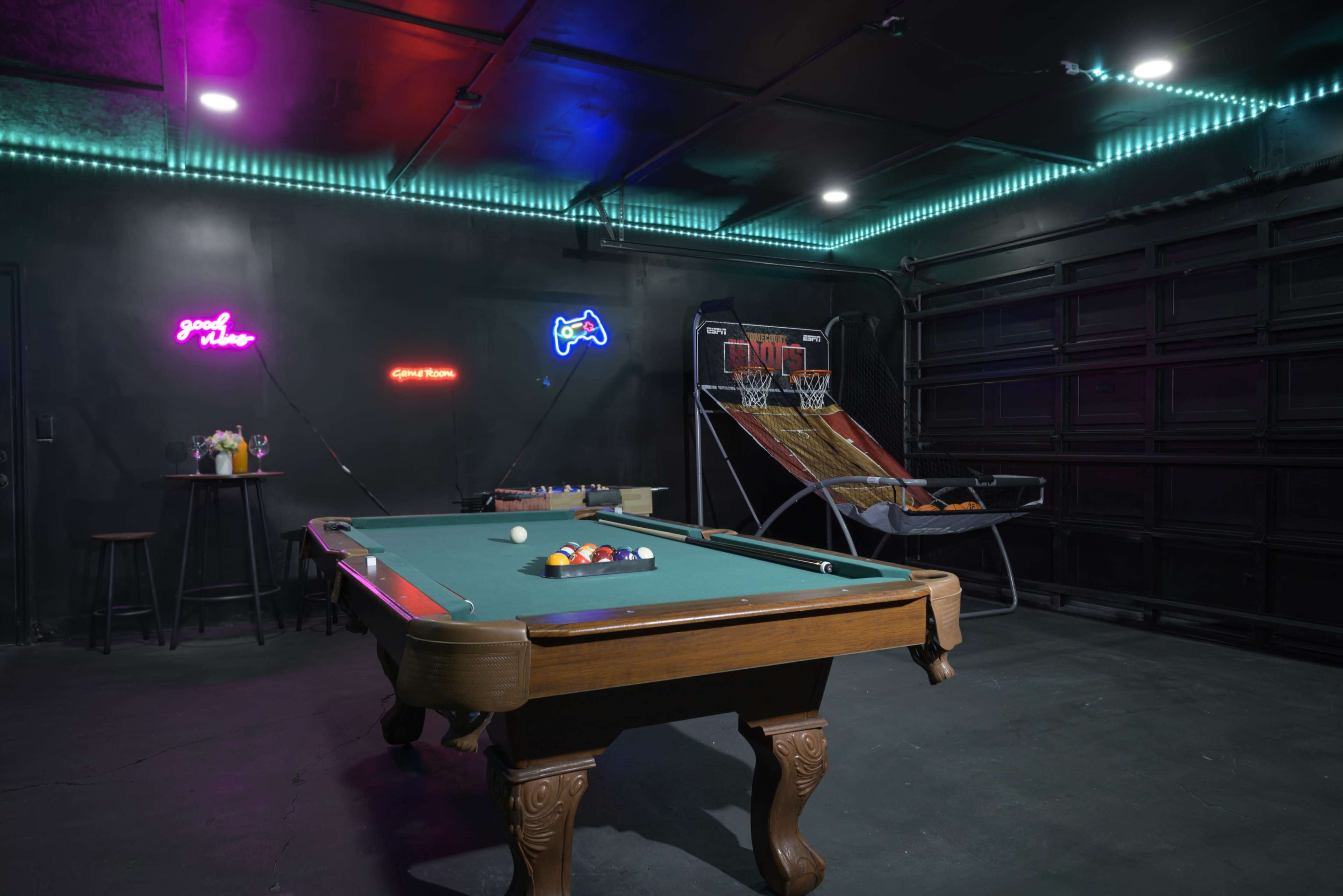 A pool table and a basketball hoop are arranged in a dimly lit game room decorated with colorful neon signs.