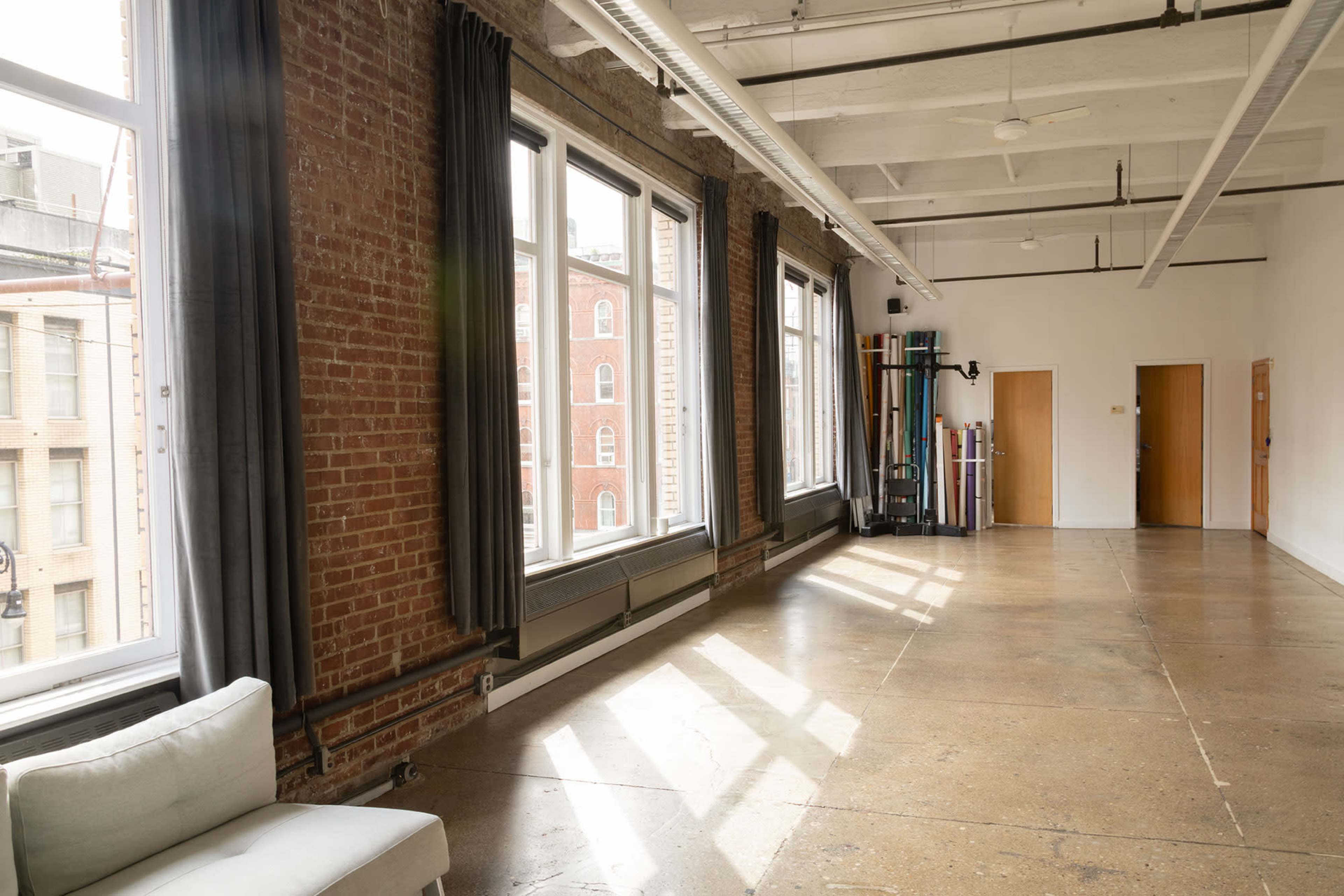 The image shows a spacious, minimalist room with large windows, exposed brick walls, and polished concrete flooring.