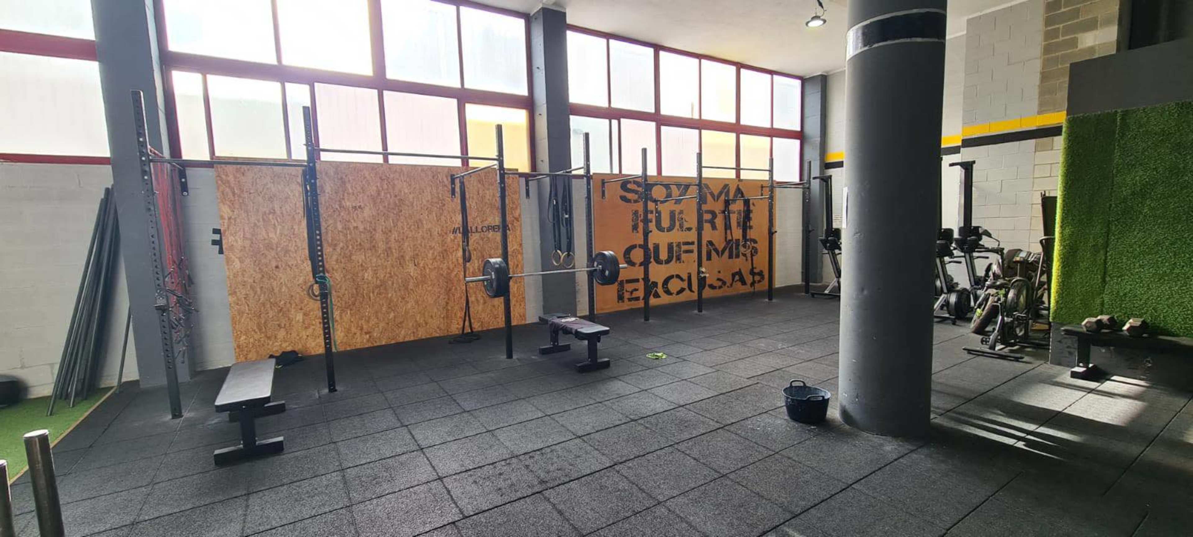 An indoor gym space featuring weightlifting equipment, benches, and a large wooden wall with text, all under bright windows.