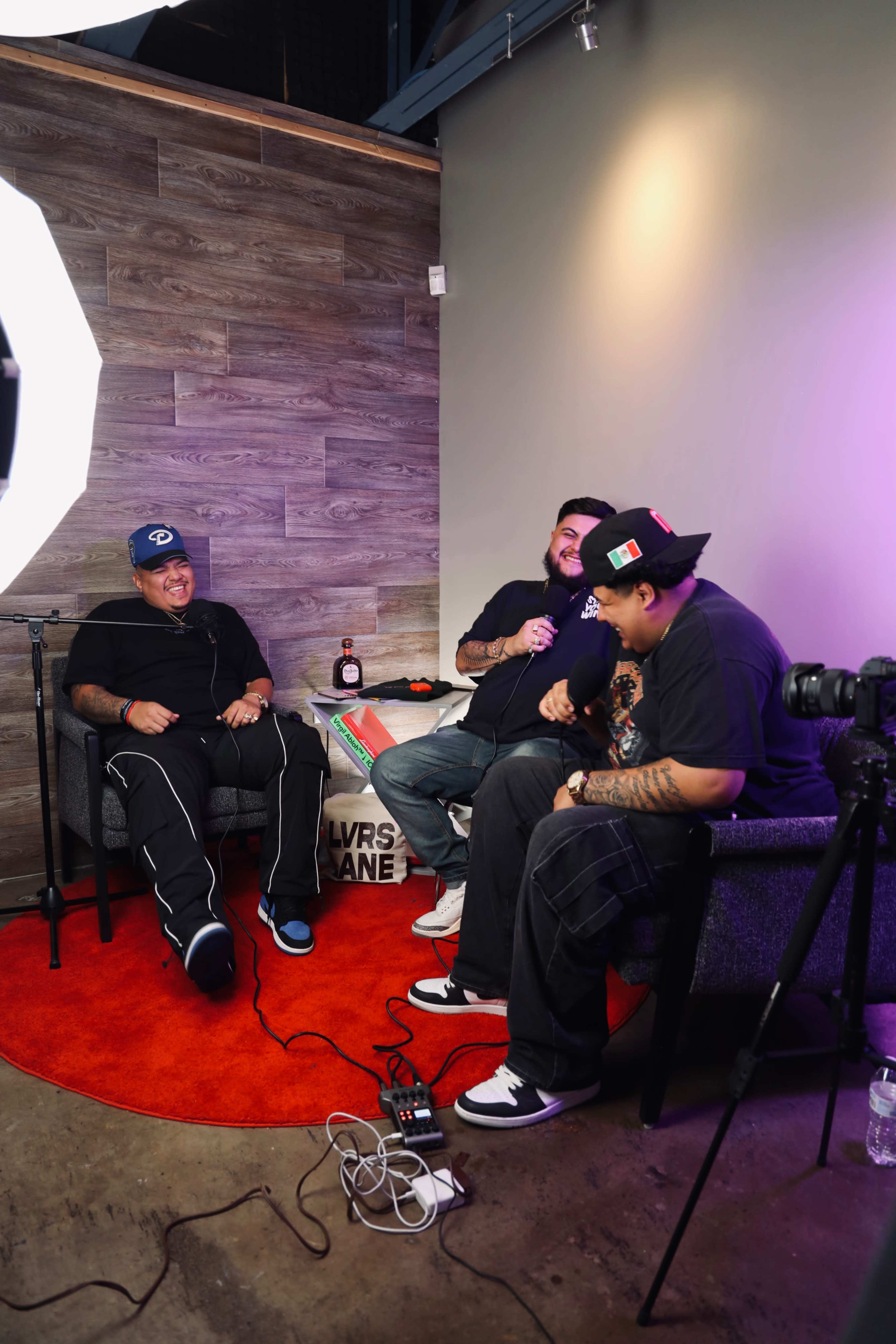Three men are sitting in a casual indoor setting, engaged in conversation, with a microphone setup, cameras, and a bottle of liquor nearby.