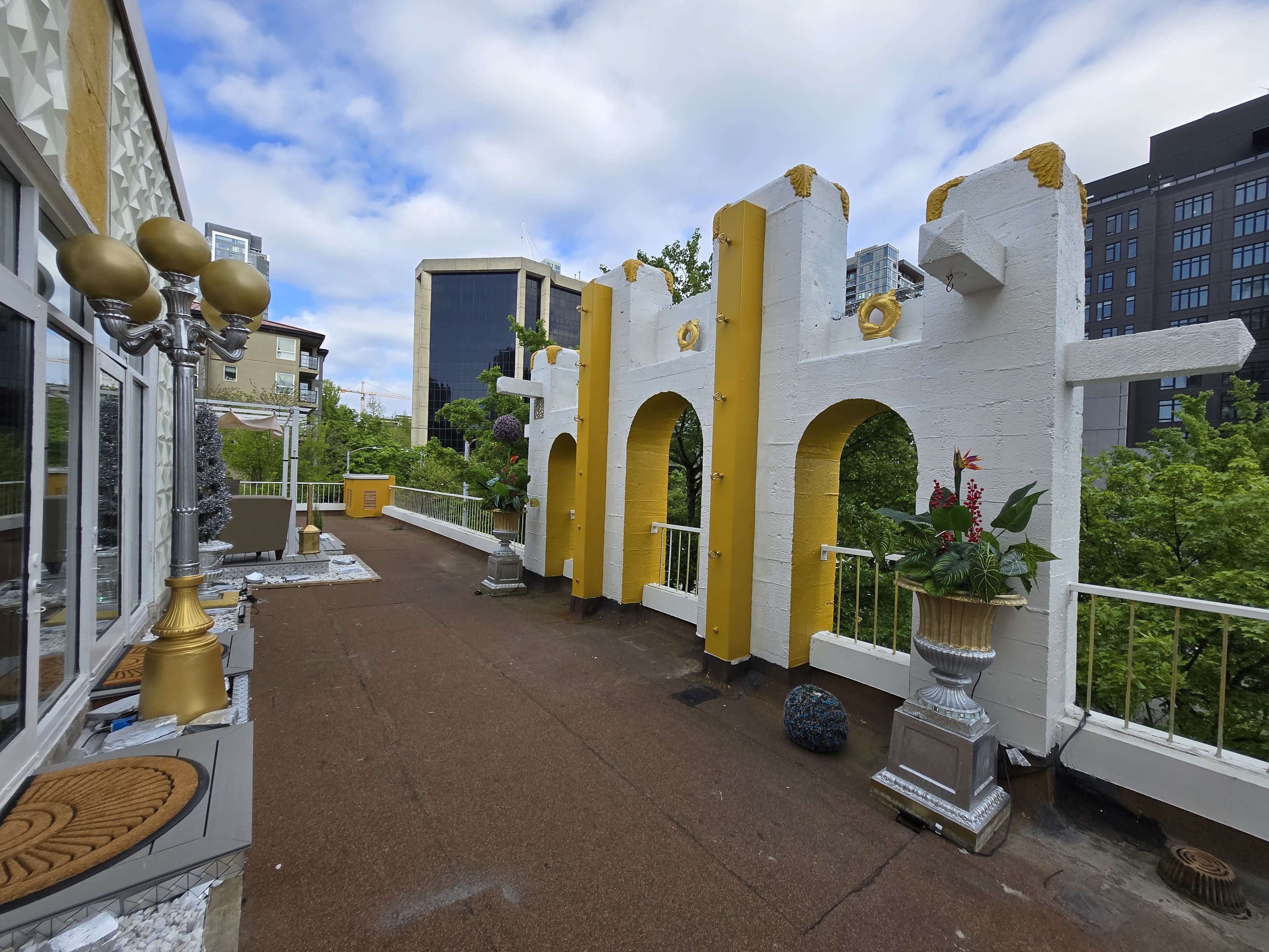 The image shows a rooftop terrace featuring white and yellow wall structures with decorative planters, flanked by large lamps and surrounded by greenery.