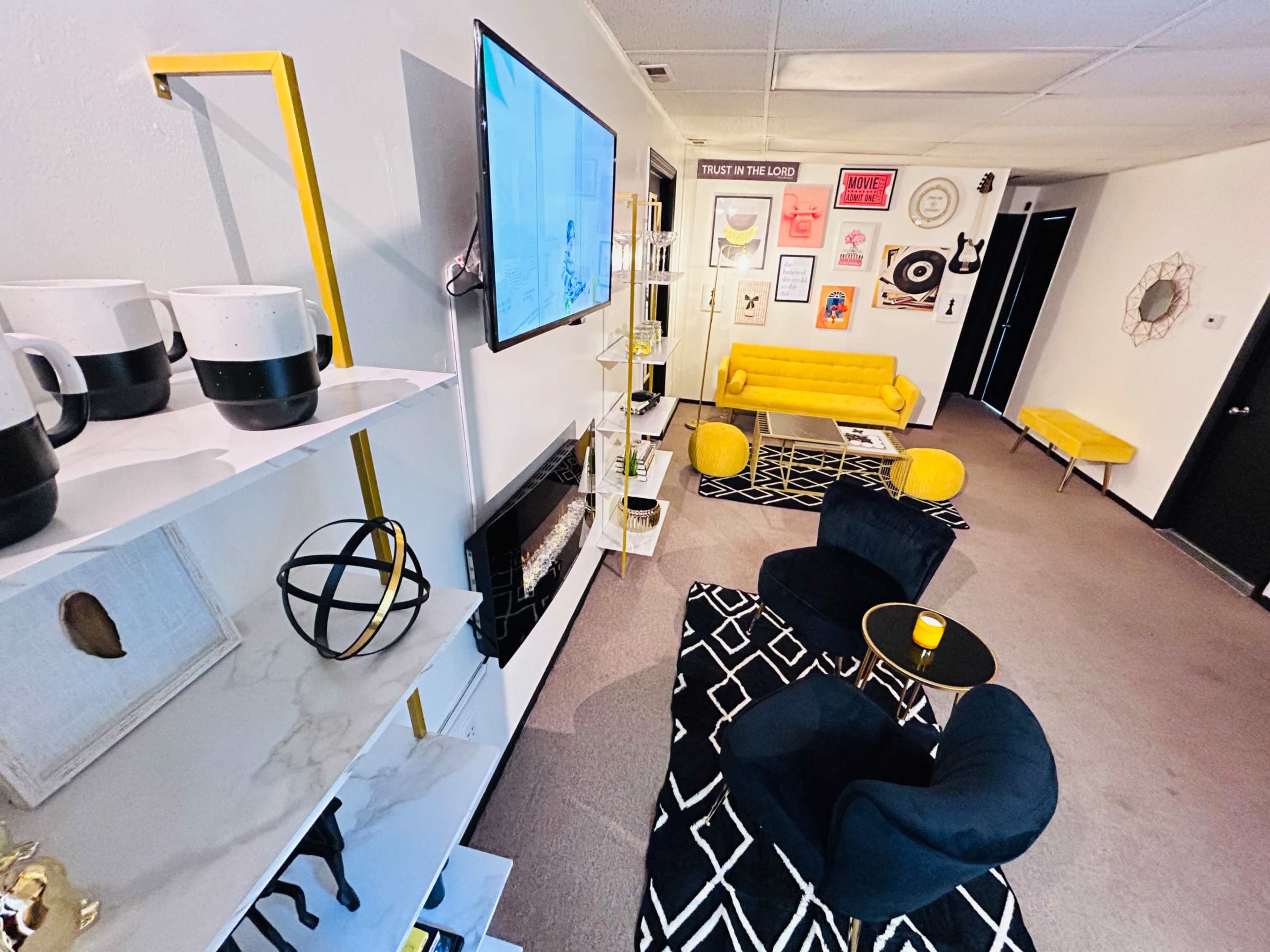 The image shows a modern living room with yellow furniture, black chairs, a wall-mounted TV, and decorative artwork on the walls.