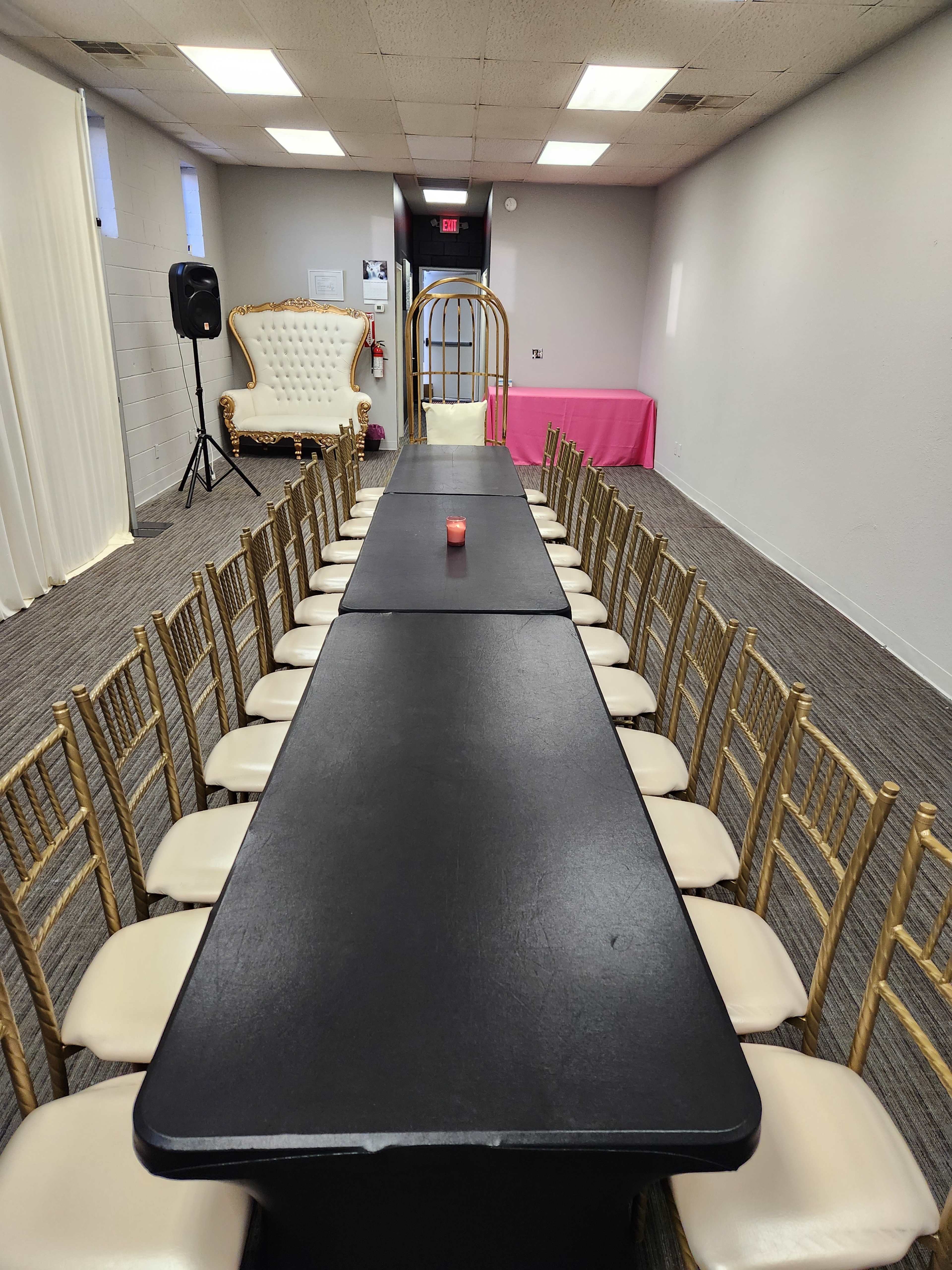 A long black table surrounded by gold chairs is set in a room with a white throne-style chair in the corner and a pink table at the far end.