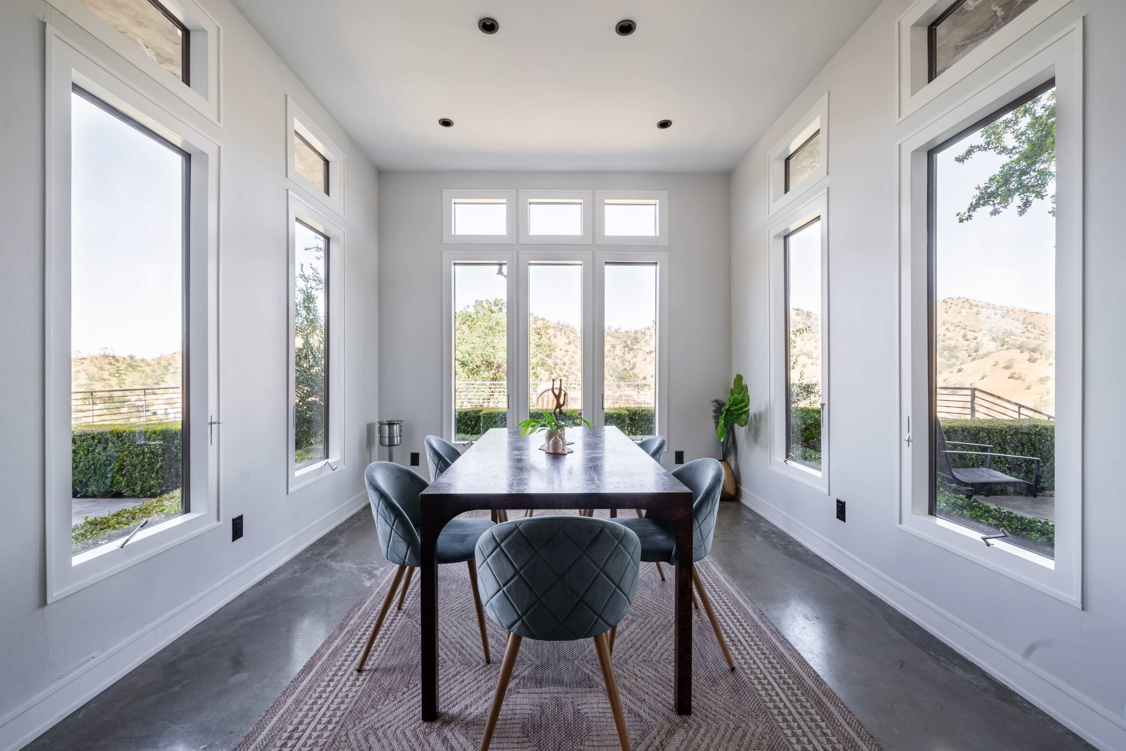 A rectangular dining table with upholstered chairs is positioned in a bright room surrounded by large windows that offer a view of a hillside.