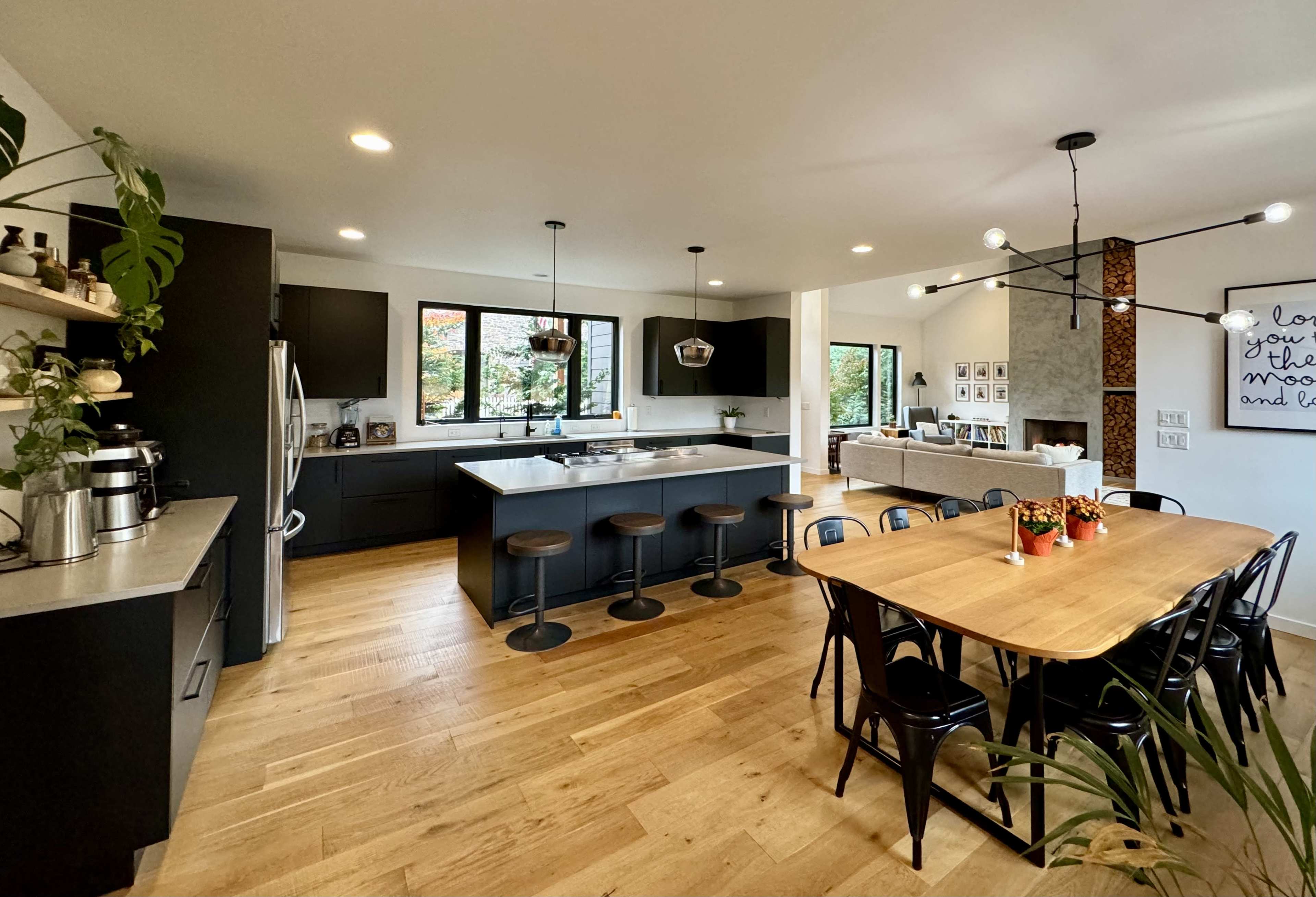 A modern kitchen features a spacious layout with a central island, bar stools, and a large dining table, complemented by hardwood flooring and large windows.