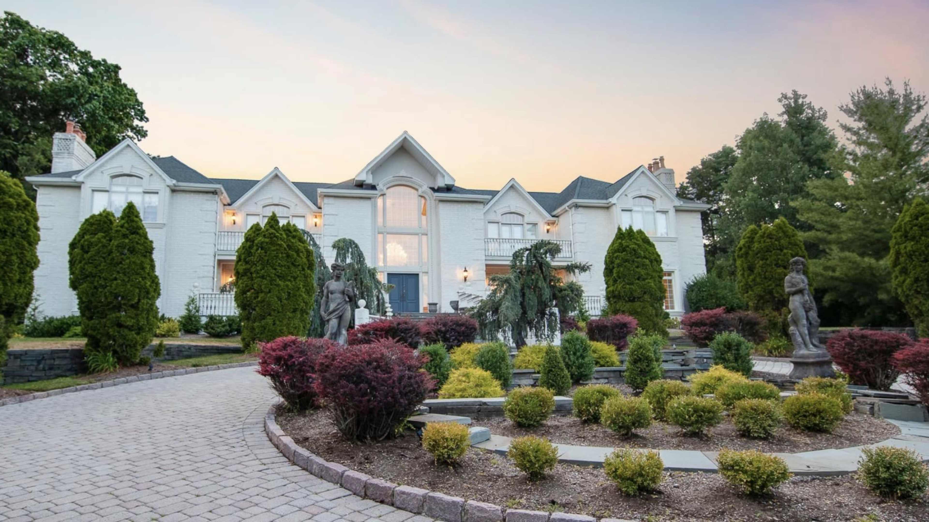 A large white mansion with a symmetrical facade is surrounded by landscaped gardens featuring shrubs and statues.