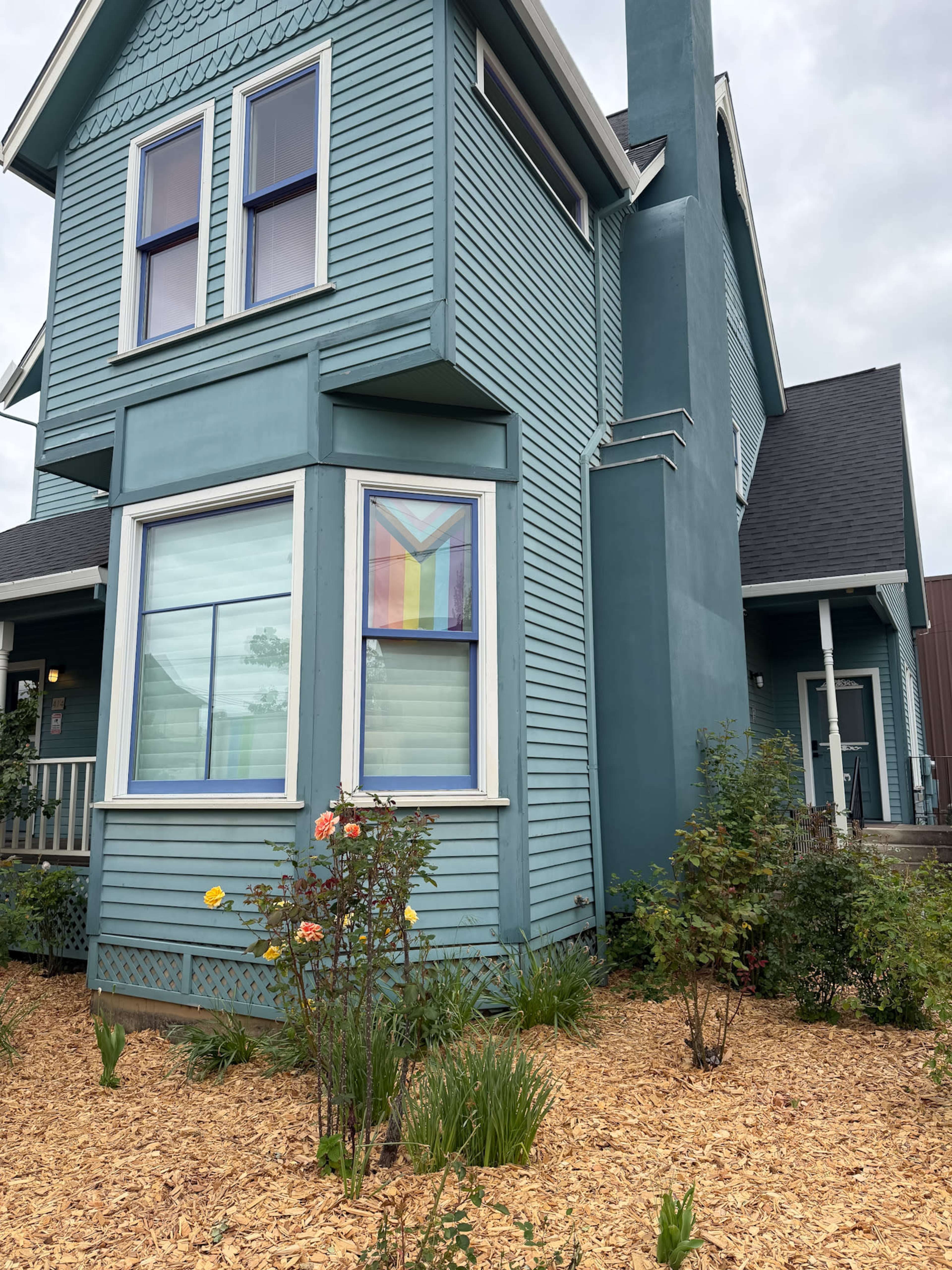 A blue house with a large window displays a rainbow flag, surrounded by a garden with flowers and mulch.