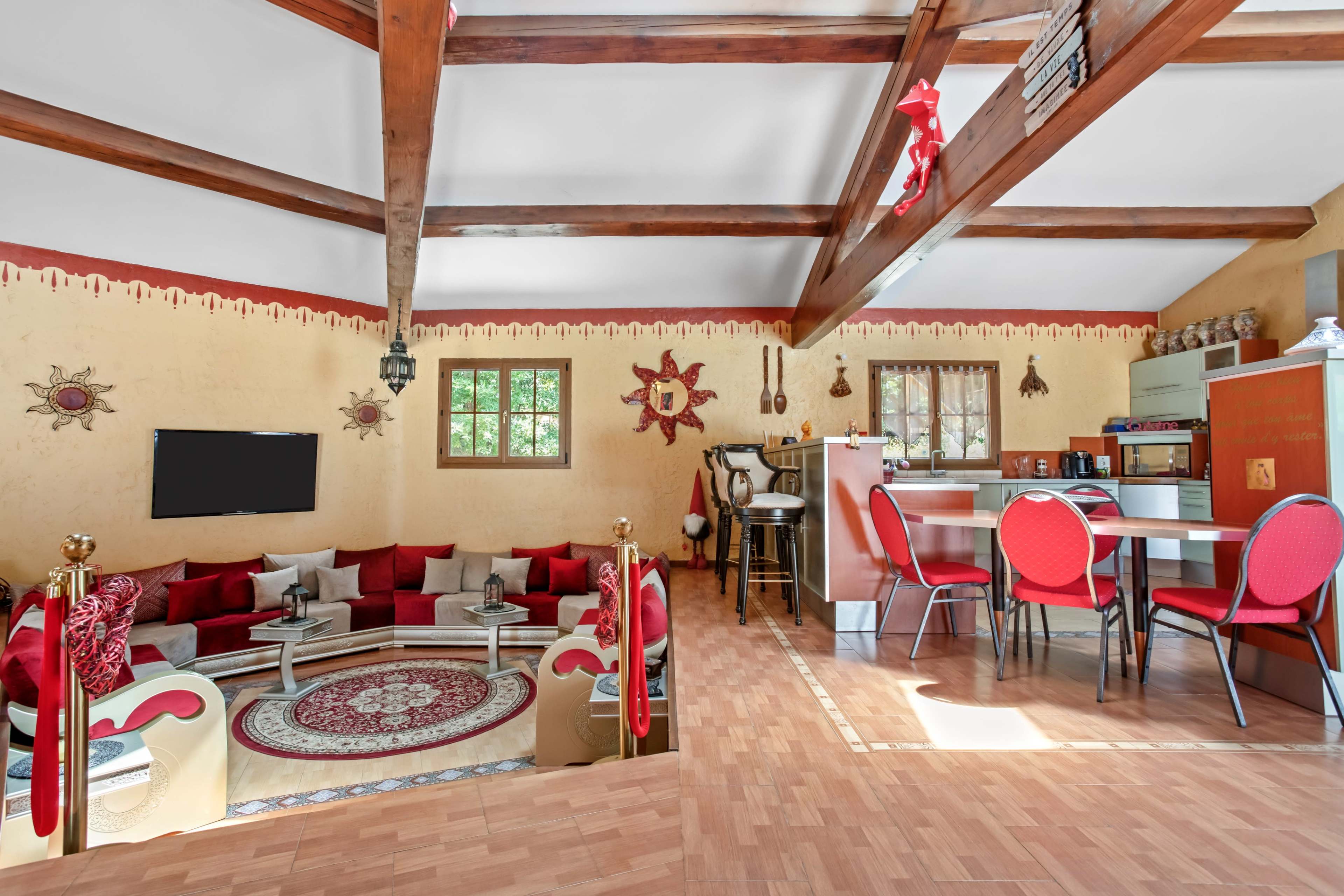 The image shows a cozy living area with a circular seating arrangement, a mounted television, and a dining area featuring red chairs and modern appliances.