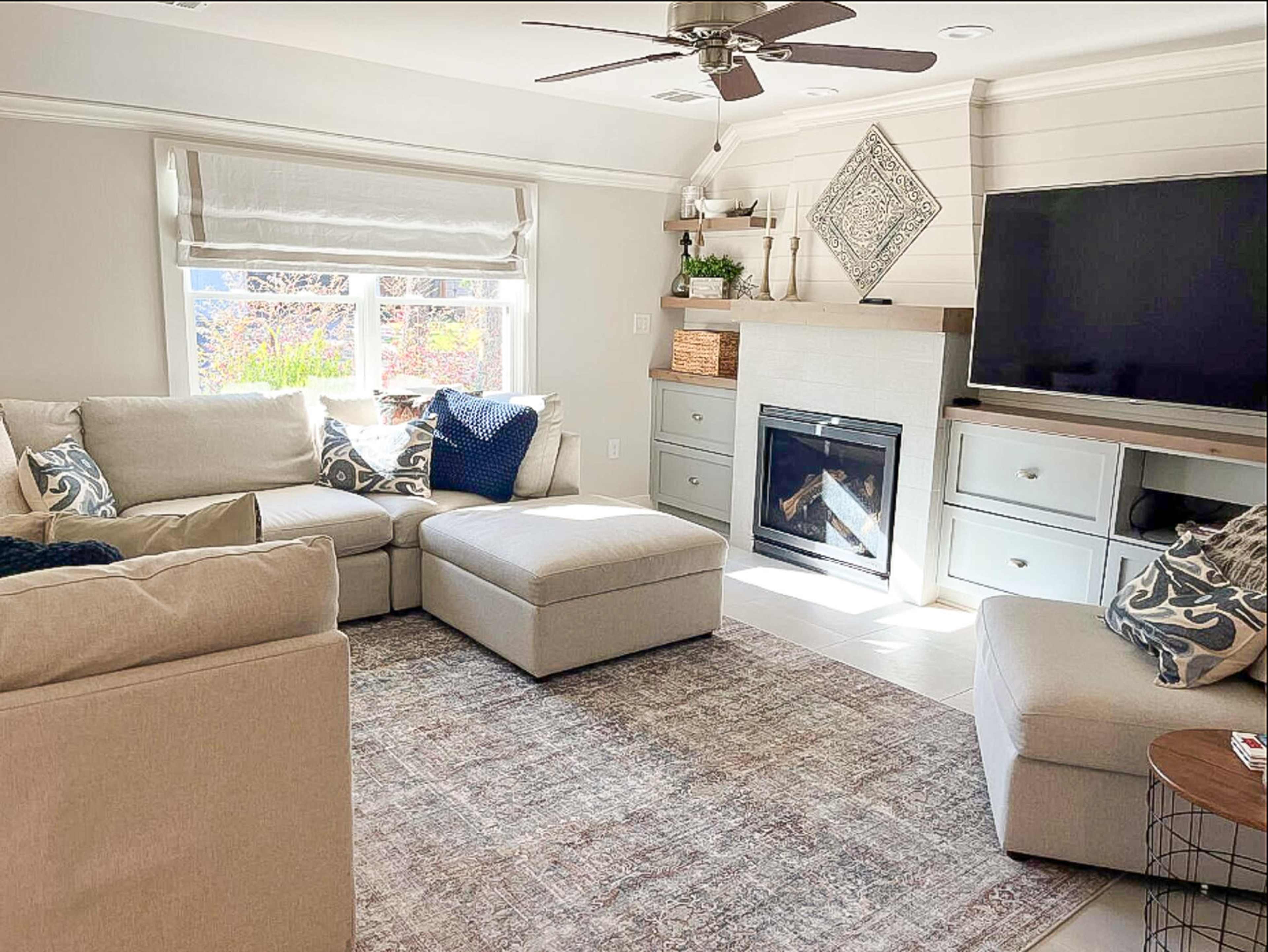 A bright living room featuring a sectional sofa, an ottoman, a fireplace, and a television on a cabinet against a light-colored wall.