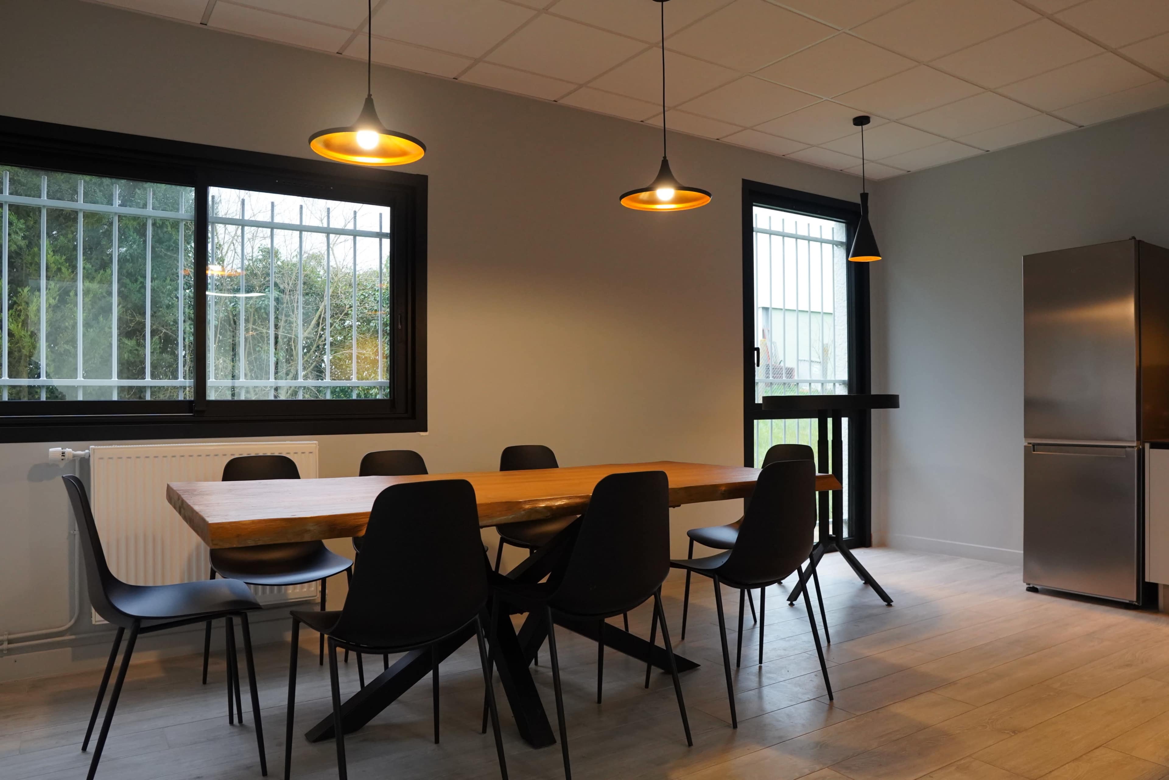 A dining area features a long wooden table surrounded by black chairs, with two hanging lights above and a refrigerator in the corner.