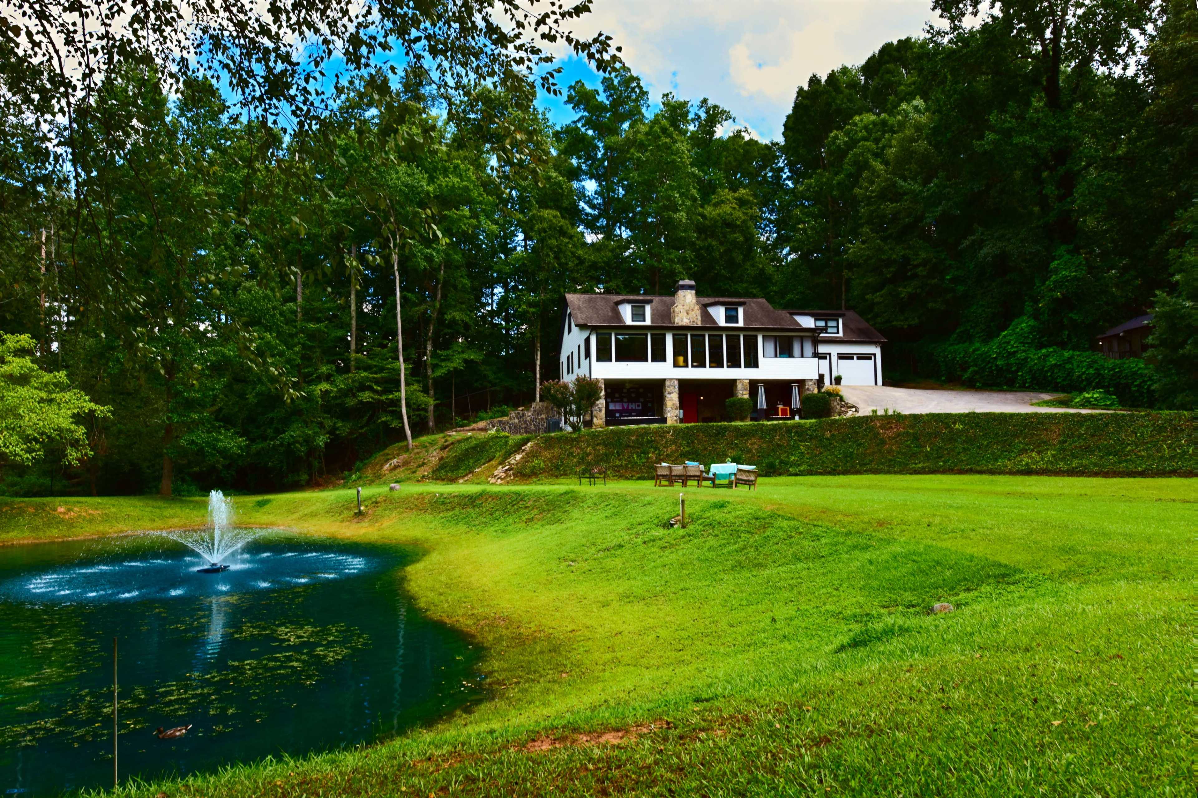 A large house situated near a pond with a fountain, surrounded by trees and a well-maintained lawn.