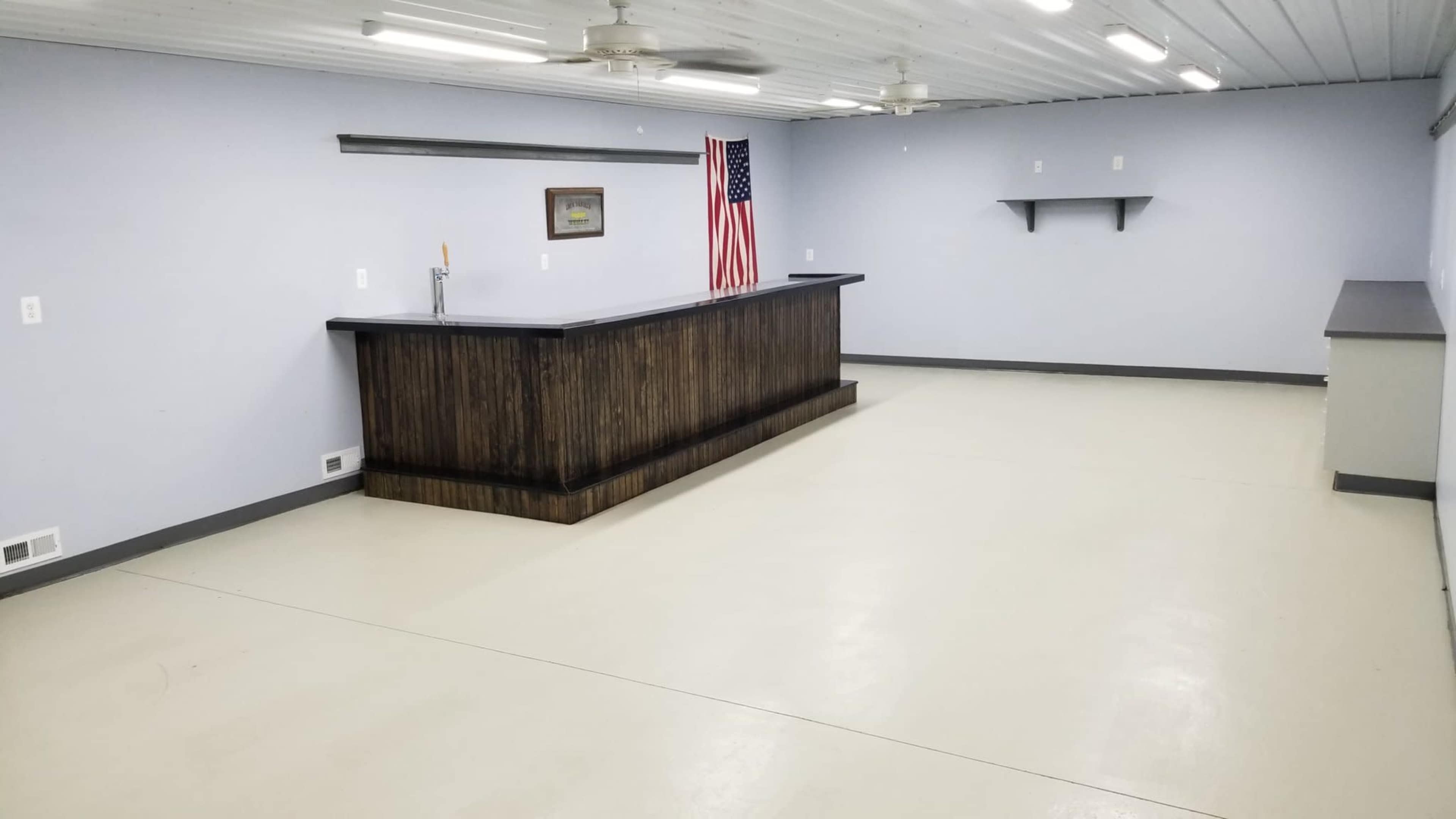 An empty room with a wooden bar at one end, a shelf on the wall, and an American flag displayed nearby.