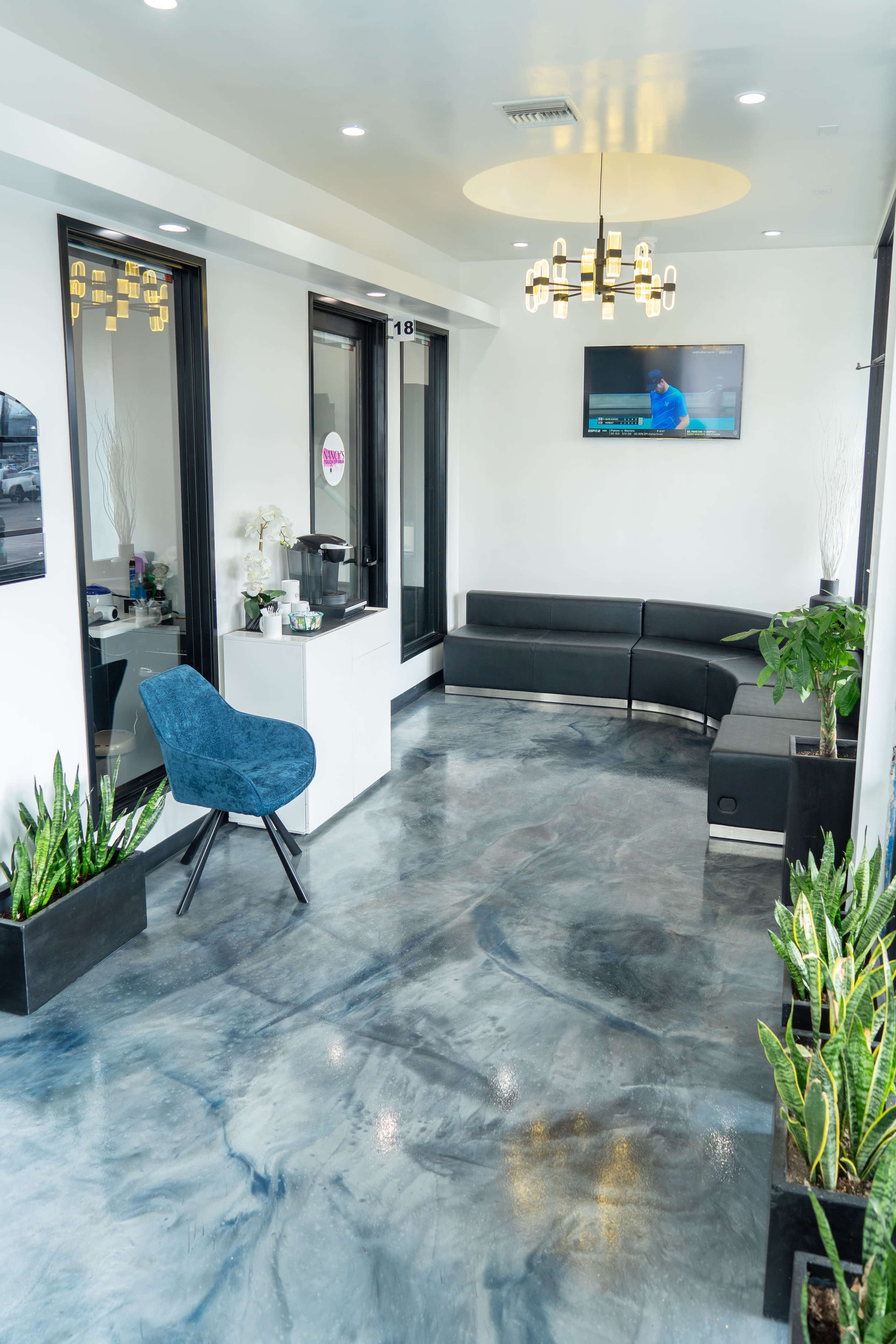 A modern reception area features sleek seating, a large TV, and potted plants against polished concrete flooring.