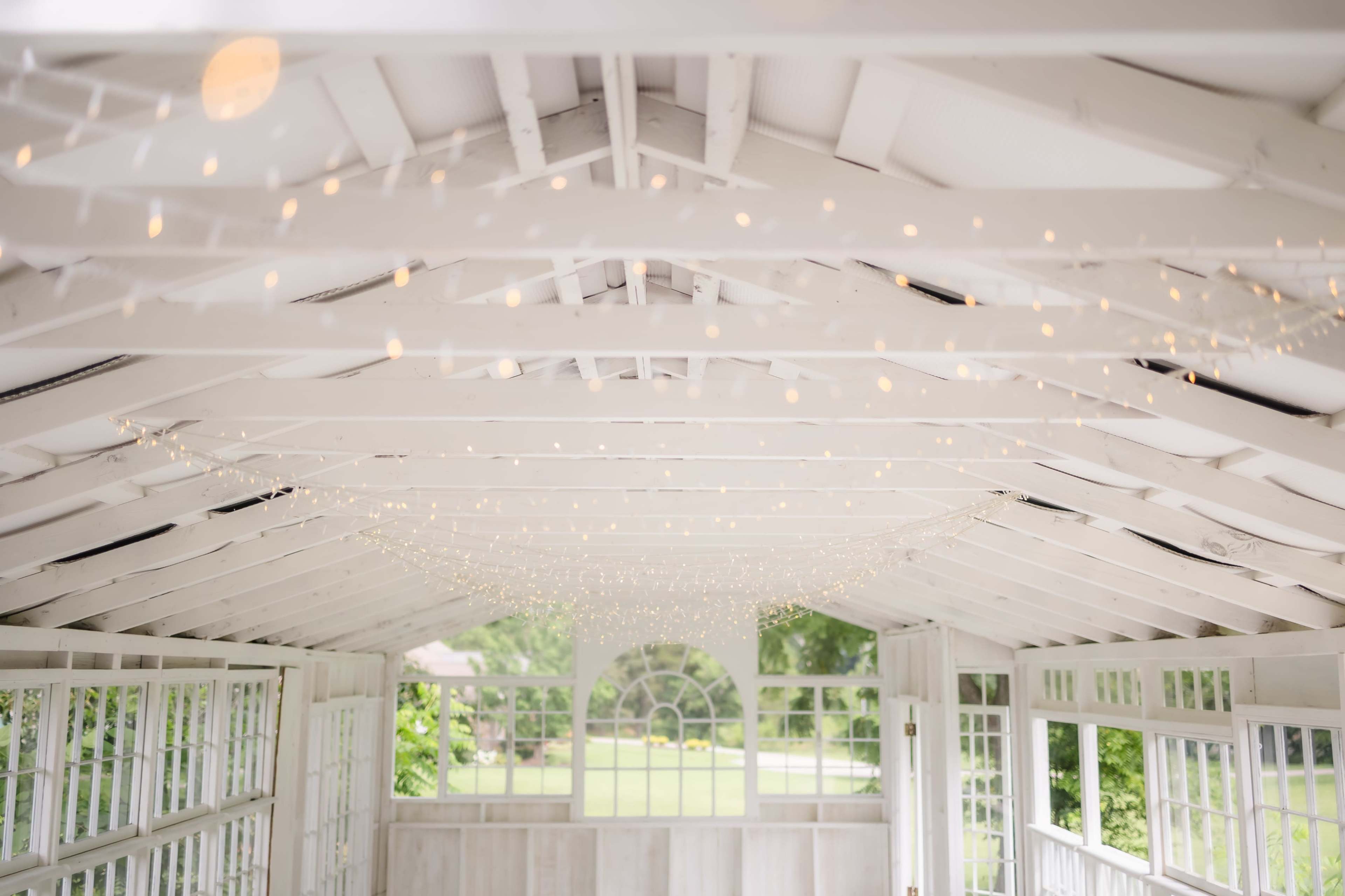 The image shows a bright, airy interior of a white-beamed structure with string lights hanging from the ceiling and large windows overlooking a green landscape.