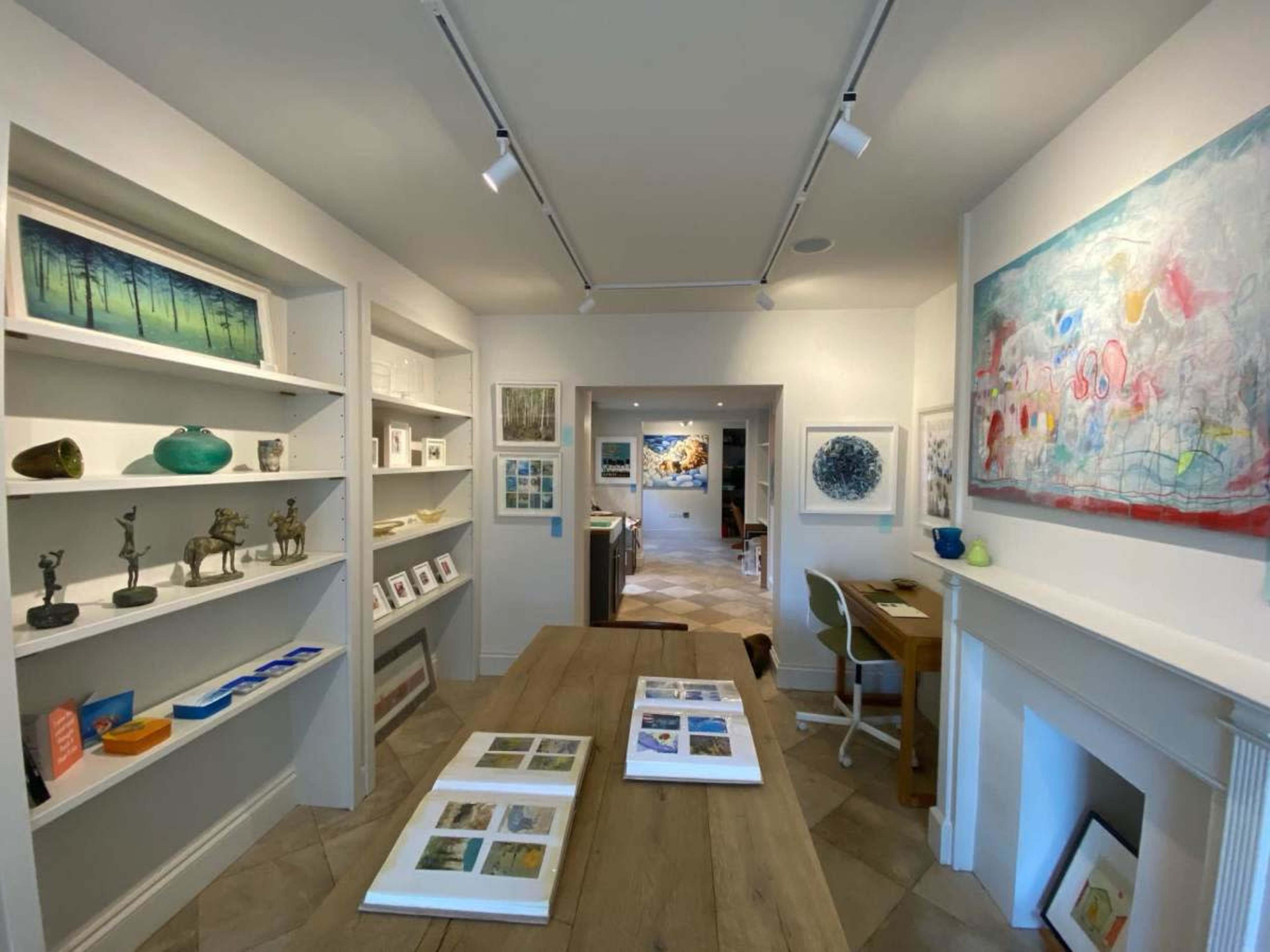 The image shows a well-lit art gallery featuring various artworks displayed on shelves and walls, with a wooden table in the foreground and a desk in an adjoining room.