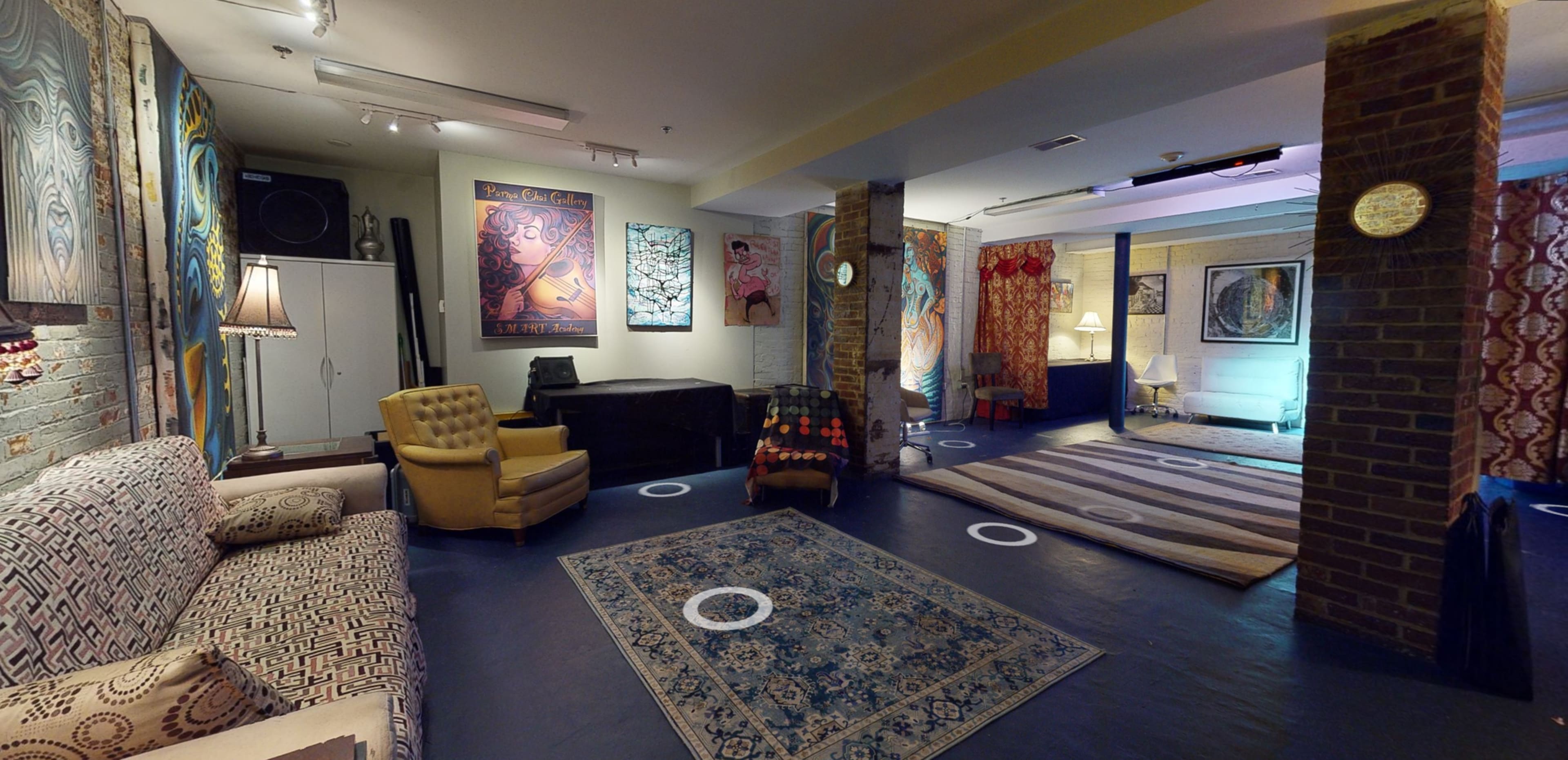 The image shows a spacious art studio or gallery with various artworks displayed on the walls, patterned rugs on the floor, and a mix of seating arrangements.