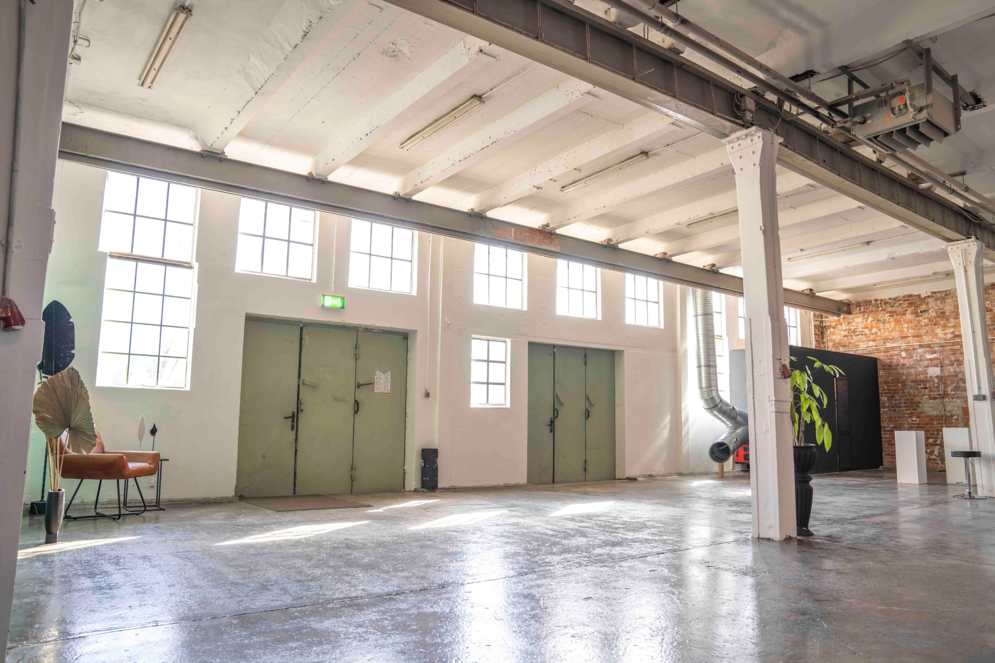 The image shows a spacious, industrial-style room with large windows, exposed brick walls, and two green double doors on one side.